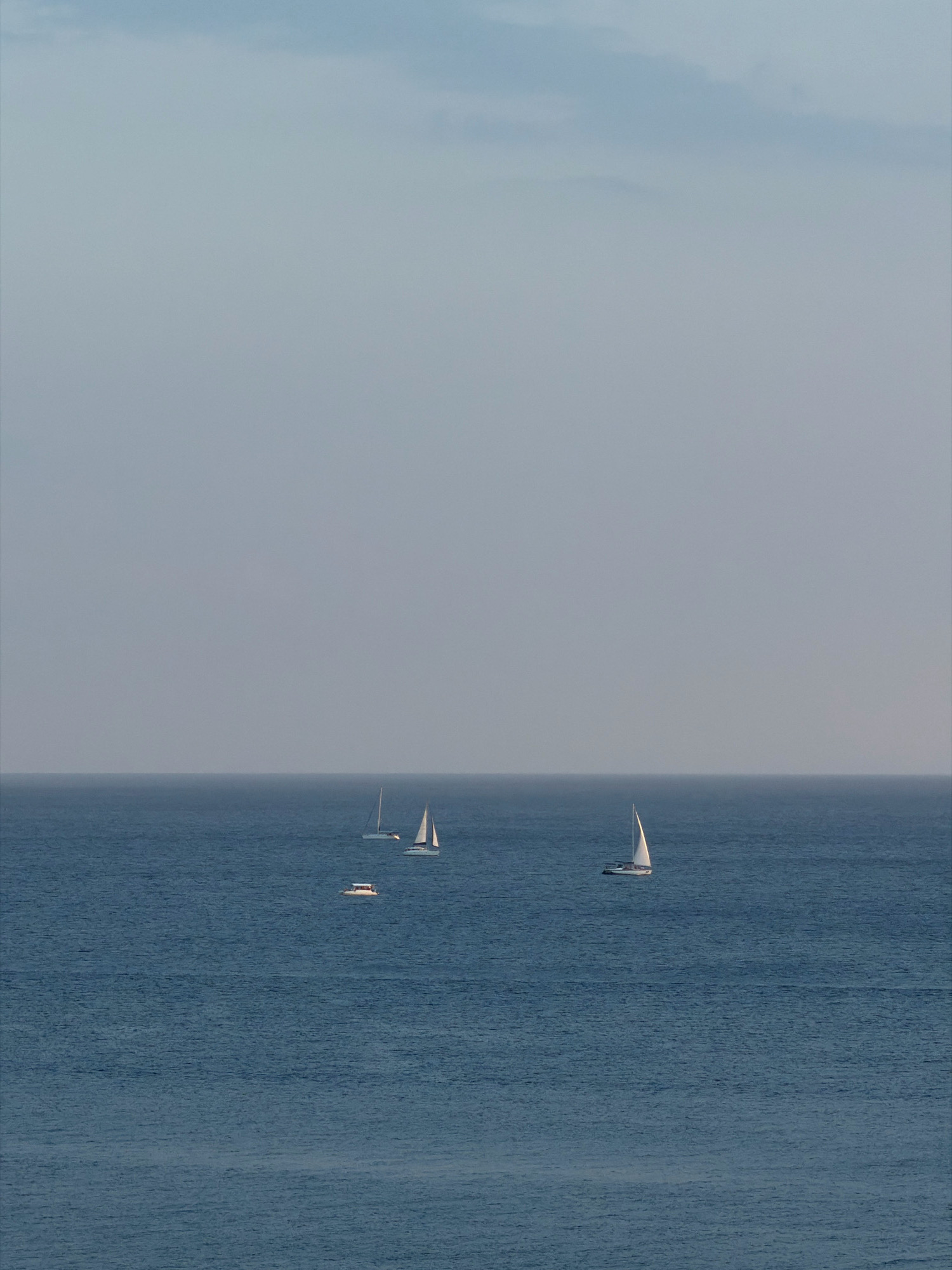 A wide-angle, high-angle shot captures a serene seascape with four vessels sailing on the calm, deep blue water. The horizon line is clearly defined, separating the water from a pale, overcast sky. The two sailboats on the right are closer to each other, with white sails catching the diffused light. A single sailboat is positioned to the left of the pair, and a smaller motorboat is even further to the left, nearer the center of the frame. The scene is devoid of any landmass or coastal features, suggesting an open sea environment. The overall impression is one of tranquility and vastness.