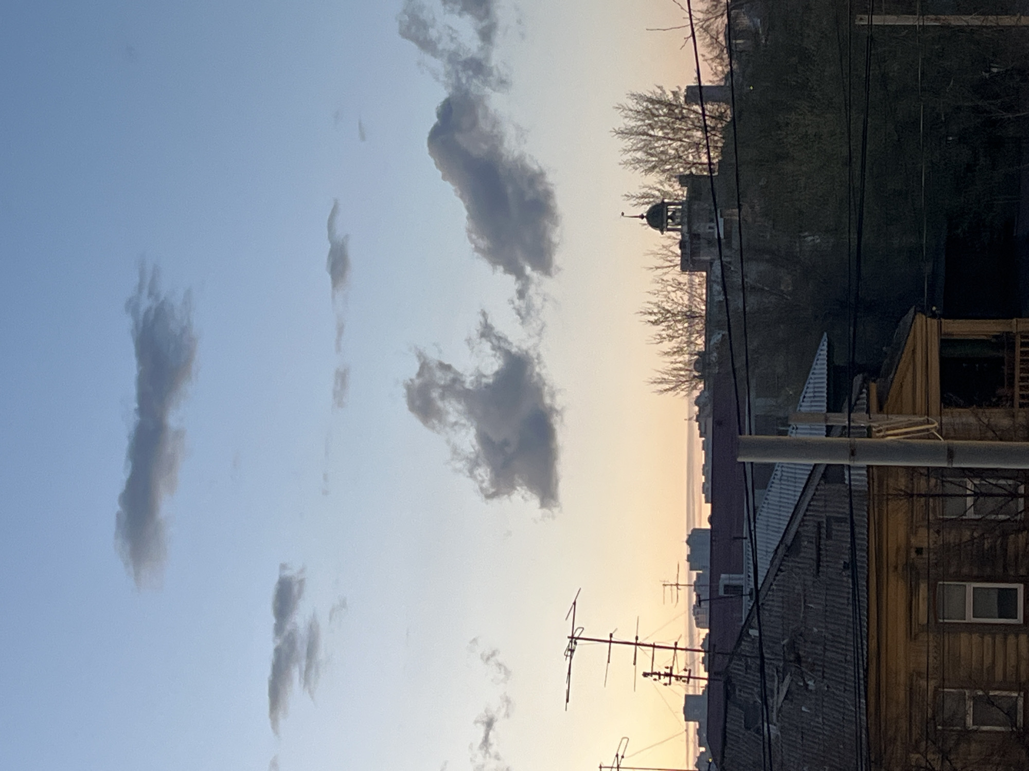 The image shows a cityscape at dusk, with a blue sky transitioning to orange near the horizon. Several elongated, gray clouds are visible in the sky. Below, rooftops of buildings are interspersed with bare tree branches and telephone poles with antennas. A church or similar building with a small dome is visible behind the trees. The foreground features weathered buildings with windows. The location is in a city, but the specific city is unknown.