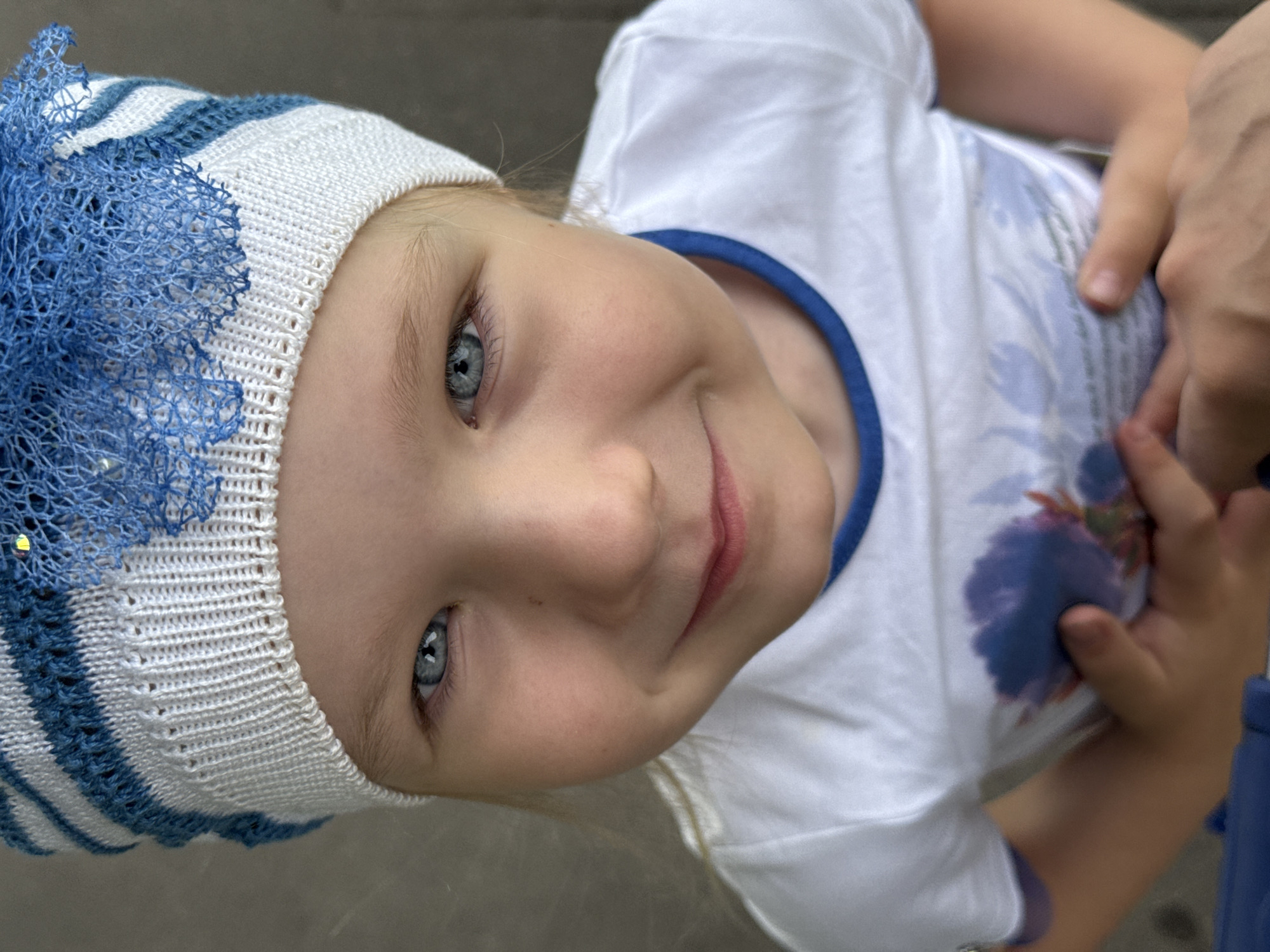 A close-up, eye-level, medium shot captures a young child's face as they look directly at the camera. The child is wearing a white t-shirt with a blue neckline and a white and blue striped knitted hat. Their eyes are a striking blue, and they have fair skin and light blonde hair peeking out from under the hat. A faint smile plays on their lips, and their hands are clasped in front of their chest. The background is a blurred, neutral gray surface, suggesting an outdoor urban environment. This image does not provide any specific details or context to determine its location within the city of Moscow, Russia.