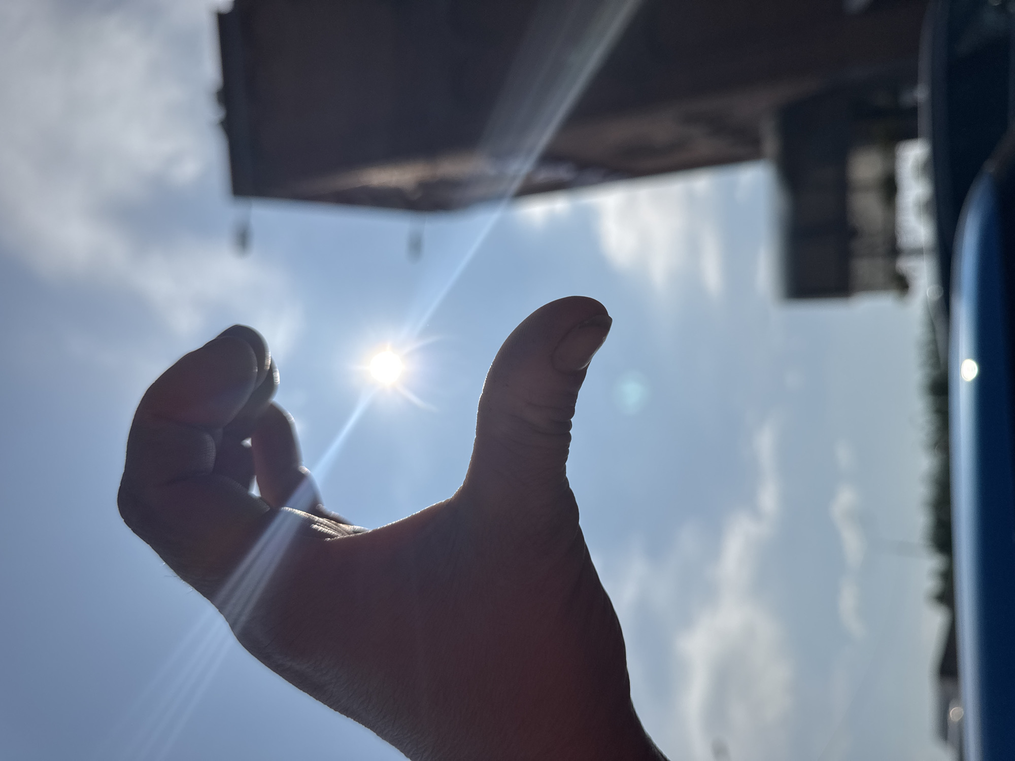 A close-up, low-angle, silhouette shot features a human hand forming a heart shape against a bright, sunlit sky. The sun, a radiant star with visible rays, is positioned within the curve of the hand. In the background, the blurry outlines of buildings and trees suggest an urban or suburban environment. The sky is predominantly blue with scattered white clouds. The image is taken outdoors during the daytime. There is no information to suggest the location is Bol'shaya Sosnova, Russia.