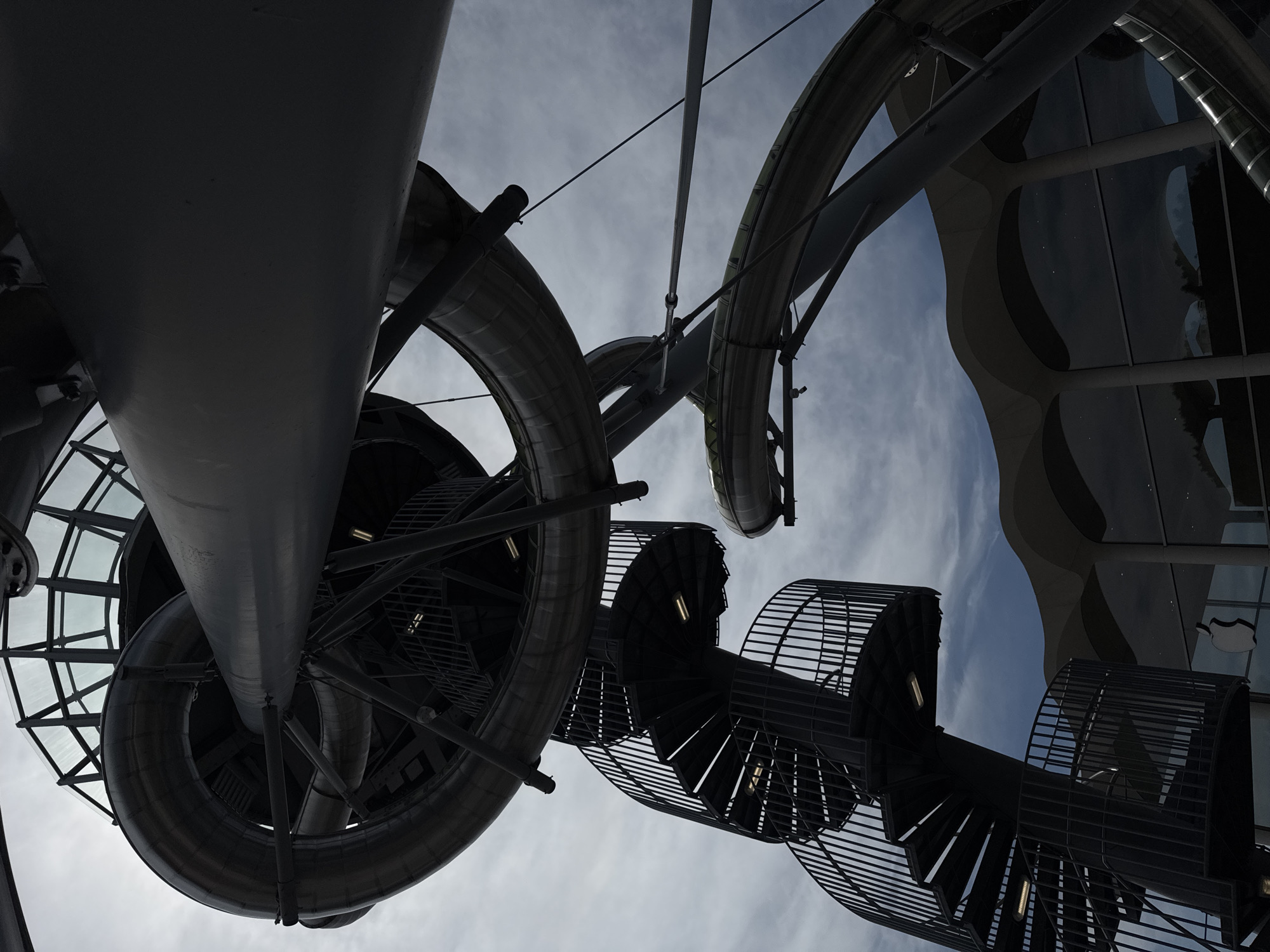 This is a low-angle shot of a large, modern outdoor art installation featuring a winding metal slide and spiral staircase in Aventura, Florida. The sculpture is predominantly gray metal with some translucent elements. The slide is a large, tubular structure that curves and twists, appearing to be part of a larger, complex design. A dark metal spiral staircase with railings is visible on the left side of the frame, ascending towards the slide structure. The sky above is cloudy, with patches of blue visible. In the background, the distinctive wavy roofline and large glass windows of a modern building, likely a retail space, are visible. An Apple logo is clearly visible on the glass facade of the building. The overall impression is one of contemporary architecture and public art.