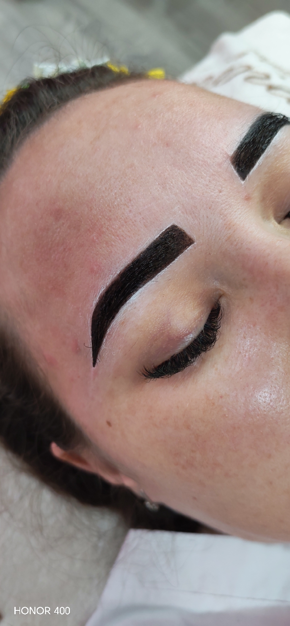Close-up image of a woman's face, specifically the right side including the eyebrow and eye, likely in Noviy, Russia. The subject is laying down. Her eyebrows are dark and heavily defined with white markings around them. The right eye is closed and has long, dark eyelashes. The skin is fair with some visible freckles and redness. The lower left corner of the image contains the text "HONOR 400". The background consists of blurry light tones.