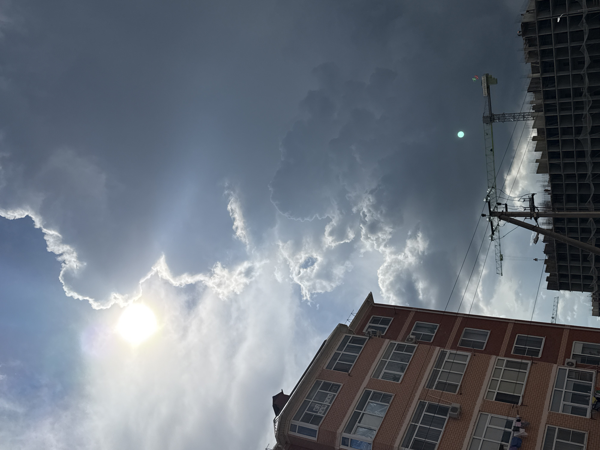 The image captures a view in an urban environment in Unknown, Unknown, dominated by a partly cloudy sky. The sun is visible on the left side of the image, partially obscured by clouds. Below the sky, there are two buildings of different construction. One is a brick building with multiple windows, positioned at a diagonal angle. The other appears to be an unfinished high-rise construction, with an exposed framework of floors and columns. Power lines and a construction crane are visible near the unfinished building.