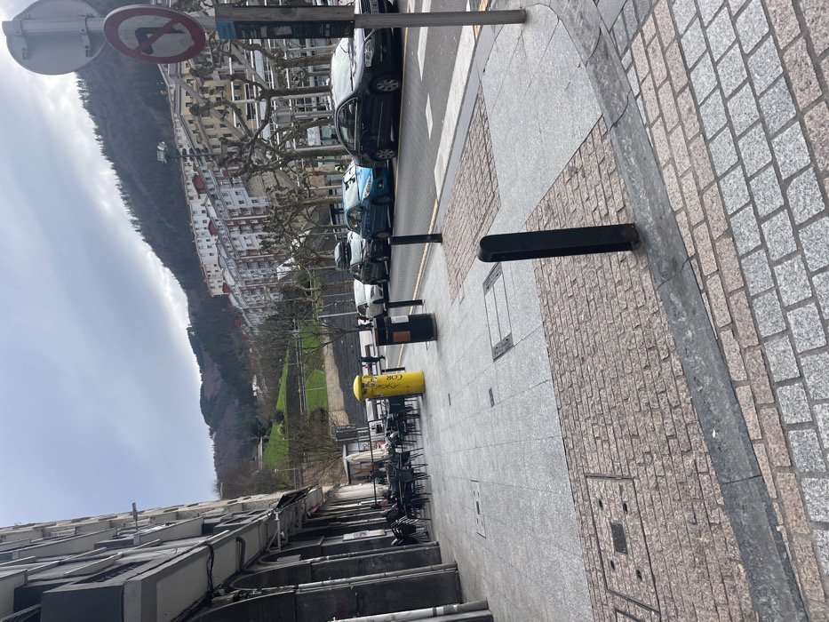 A street scene features parked cars lining the road, with a sidewalk on the right constructed with a pattern of large, light-colored bricks. A yellow cylindrical structure is positioned alongside chairs and tables, possibly part of an outdoor café area. A backdrop reveals a mountain range and buildings, set against a cloudy sky. Street signs are mounted on poles.