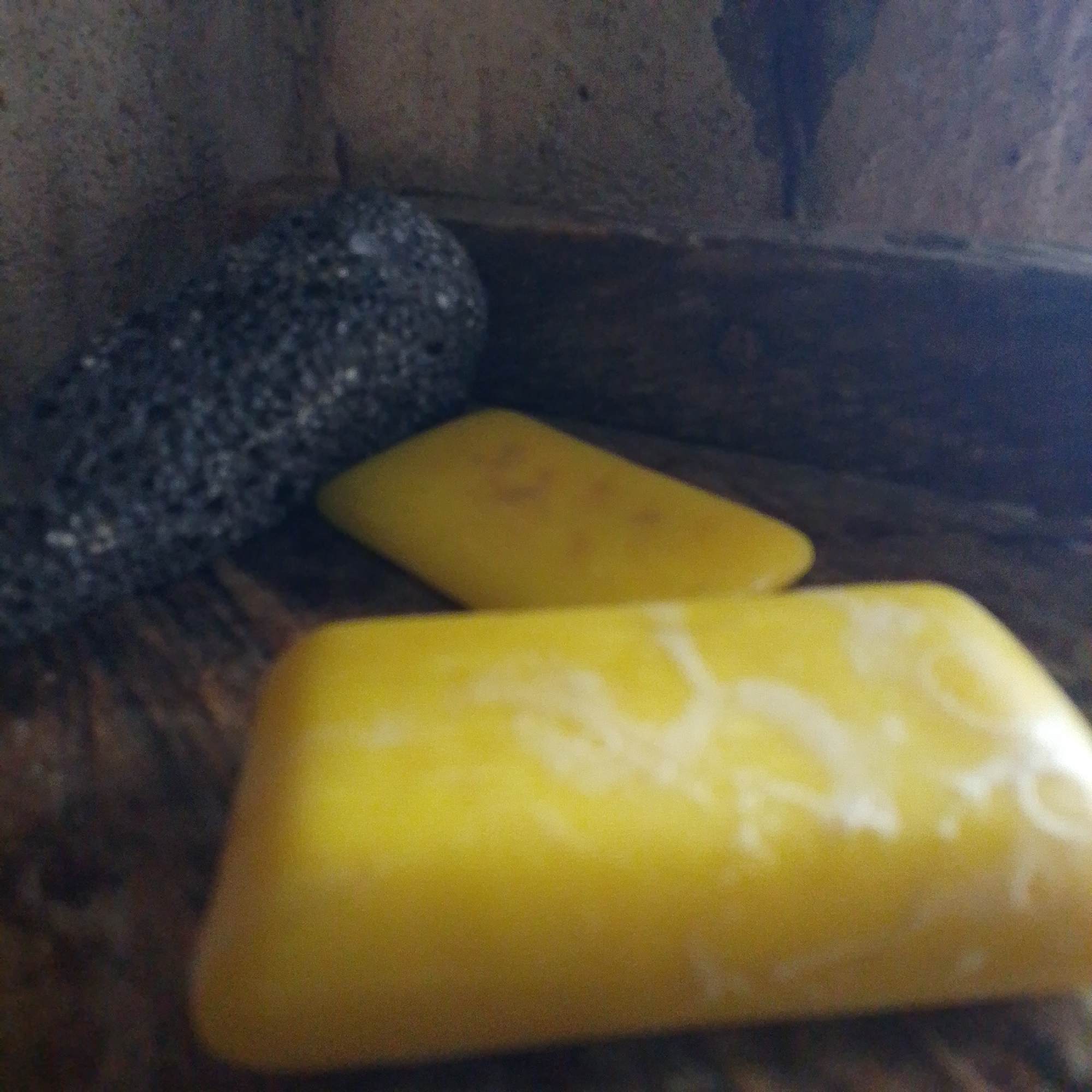 The image depicts a close-up view of two rectangular yellow objects, possibly bars of soap or wax, resting on a dark, textured surface. One object is in the foreground, slightly out of focus, with swirling lighter patterns on its surface. The second object is positioned behind and to the left of the first, also yellow, but with a less distinct texture. To the far left, partially visible, is a dark, granular object, possibly a pumice stone or a similar abrasive tool. The background is dark and out of focus, appearing to be a rough, possibly stone or wooden surface, with hints of a dark, textured wall. The scene is dimly lit, suggesting an indoor or shaded environment. No people or actions are visible. The image does not contain any information that would indicate its location in Jalingo, Nigeria.