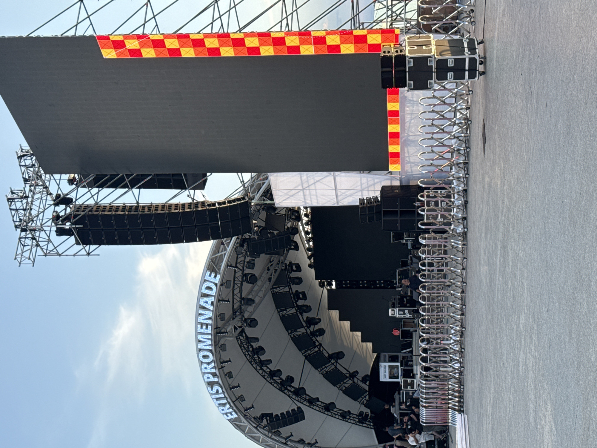 An outdoor stage setup is visible under a clear blue sky with wispy clouds. The stage features a large, dark LED screen on the right, bordered by a checkered pattern of red and yellow lights. To the left, a curved white canopy with "ERTIS PROMENADE" illuminated in blue letters is visible. Large speaker stacks are mounted on metal truss structures on either side of the stage. In front of the stage, a row of silver retractable barriers on wheels sections off the performance area. Various audio equipment, including speaker cabinets and road cases, are present on the ground. Several people are seen on and around the stage, some appearing to be setting up or managing equipment. The ground in the foreground is paved. The image depicts preparations for an event at the Ertis Promenade in Pavlodar, Kazakhstan.