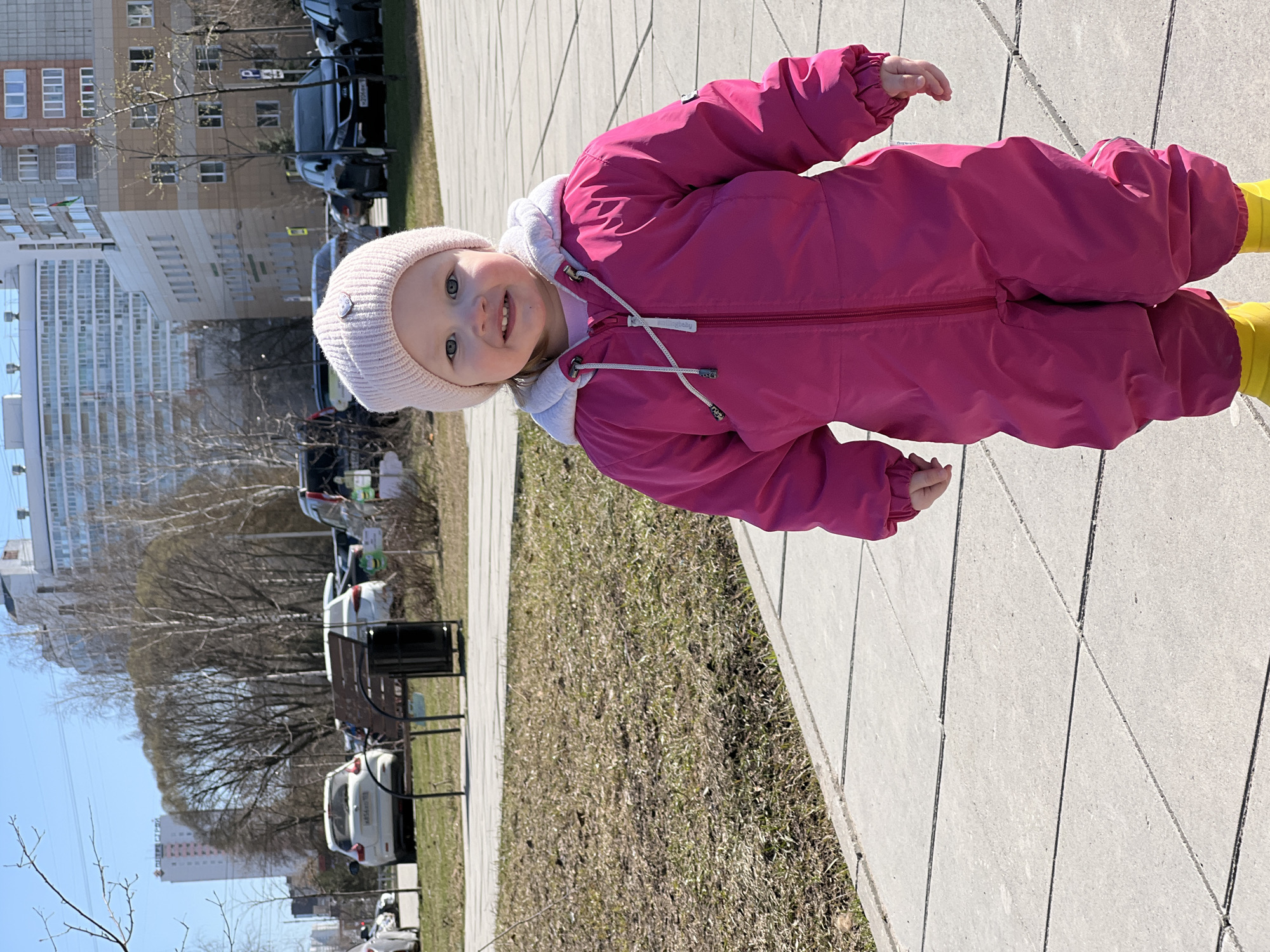 A young child in a pink snowsuit and yellow boots stands on a light gray, textured concrete sidewalk. The child is facing the viewer and smiling, wearing a light pink knit hat. The background features a grassy area with sparse, dry vegetation, a park bench, and parked cars. In the distance, tall buildings are visible under a clear blue sky. The location is Perm, Russia.