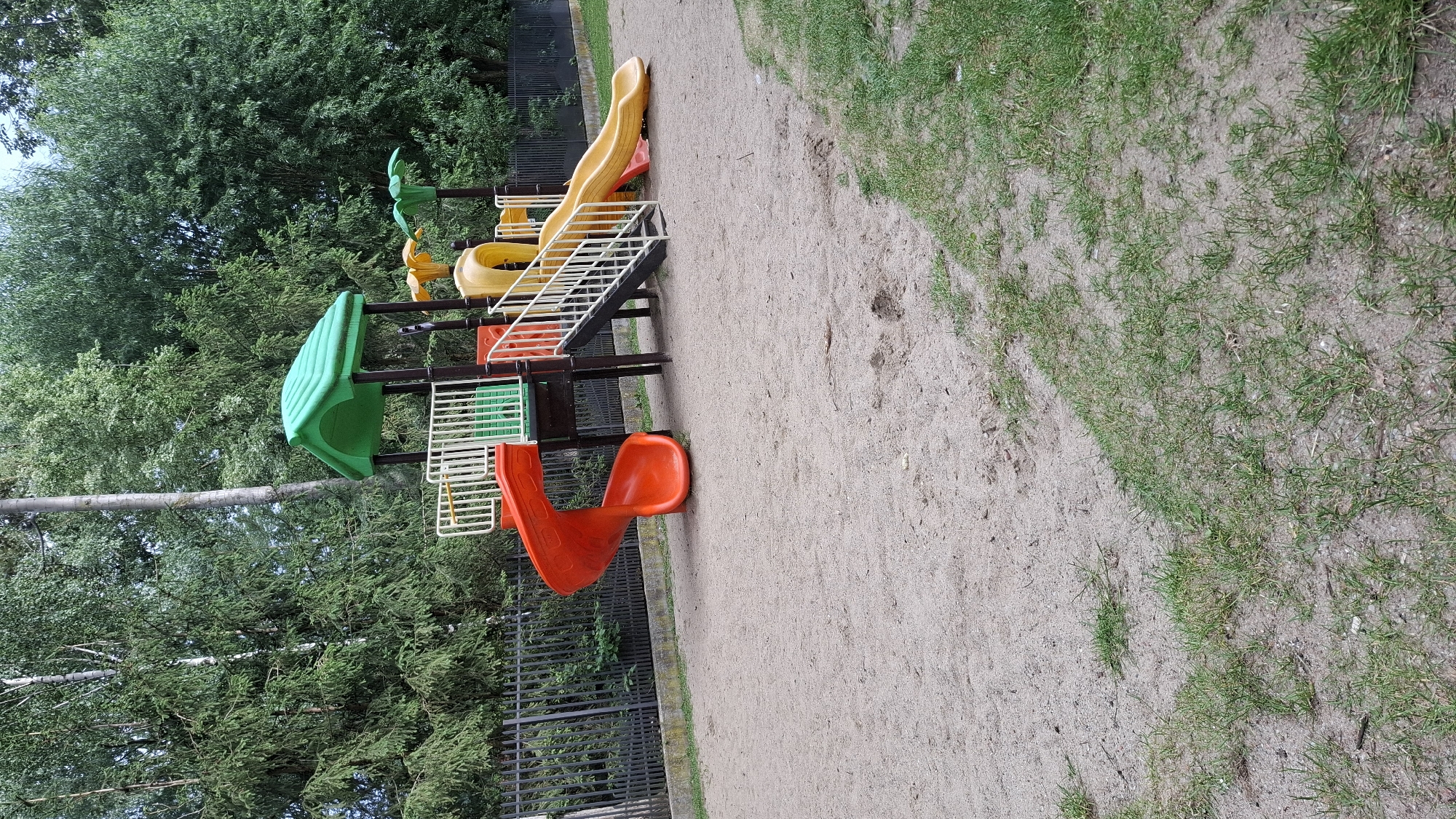 The image depicts a section of a playground with a multi-level play structure. The structure features bright plastic components in green, yellow, and orange, including slides and platforms. A yellow, wavy slide is visible at the top, and a red, curved slide is at the bottom. White stairs with black railings connect different levels of the play equipment. The playground is situated on a sandy ground with patches of green grass. In the background, a dense cluster of green trees and a dark metal fence are present. The lighting suggests it is daytime, likely with overcast conditions given the absence of strong shadows. The scene appears calm and unoccupied, implying no specific activity or event is taking place at the moment the photo was taken. There is no visible text in the image. The location context provided is Grójec, Poland.