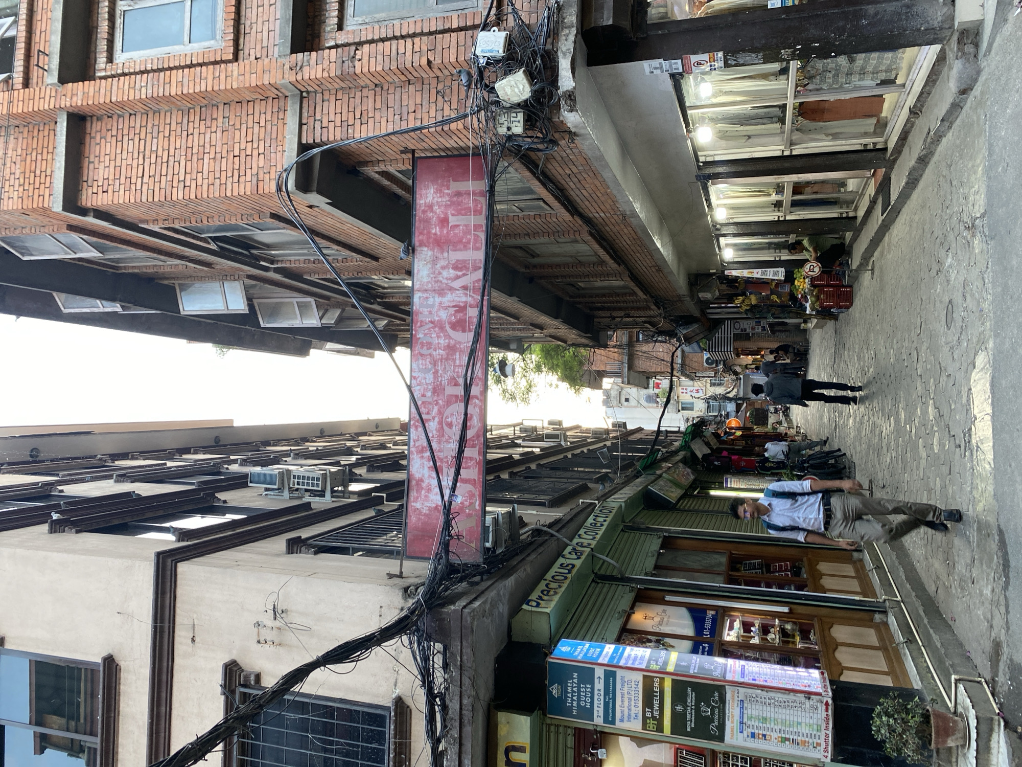 The image depicts a narrow, cobblestone street in Kathmandu Metropolitan City, Nepal, flanked by multi-story buildings on both sides. The architecture on the left features a beige facade with dark brown trim and barred windows, while the building on the right is constructed of red brick with numerous windows, some of which are open. A large, weathered red sign with white text is suspended across the street between the two buildings. Numerous electrical wires and conduits are strung haphazardly between the buildings, contributing to a cluttered overhead view.
On the left side of the street, businesses are visible, including one with a sign indicating "THAMEL HIMALAYAN GUEST HOUSE" and "Precious Care Collection." Displays of jewelry and currency exchange rates are present. A man in a white shirt and light-colored pants, carrying a blue backpack, walks away from the camera down the street.
On the right side of the street, shops with illuminated storefronts display clothing on racks. A person in a green shirt is seated on the curb in front of one of these shops. Further down the street, two individuals are walking away from the camera, and other figures are visible in the distance. The overall scene suggests a bustling urban environment, typical of commercial areas in Kathmandu. The presence of air conditioning units mounted on the buildings and the style of the signage further place the location within the city.