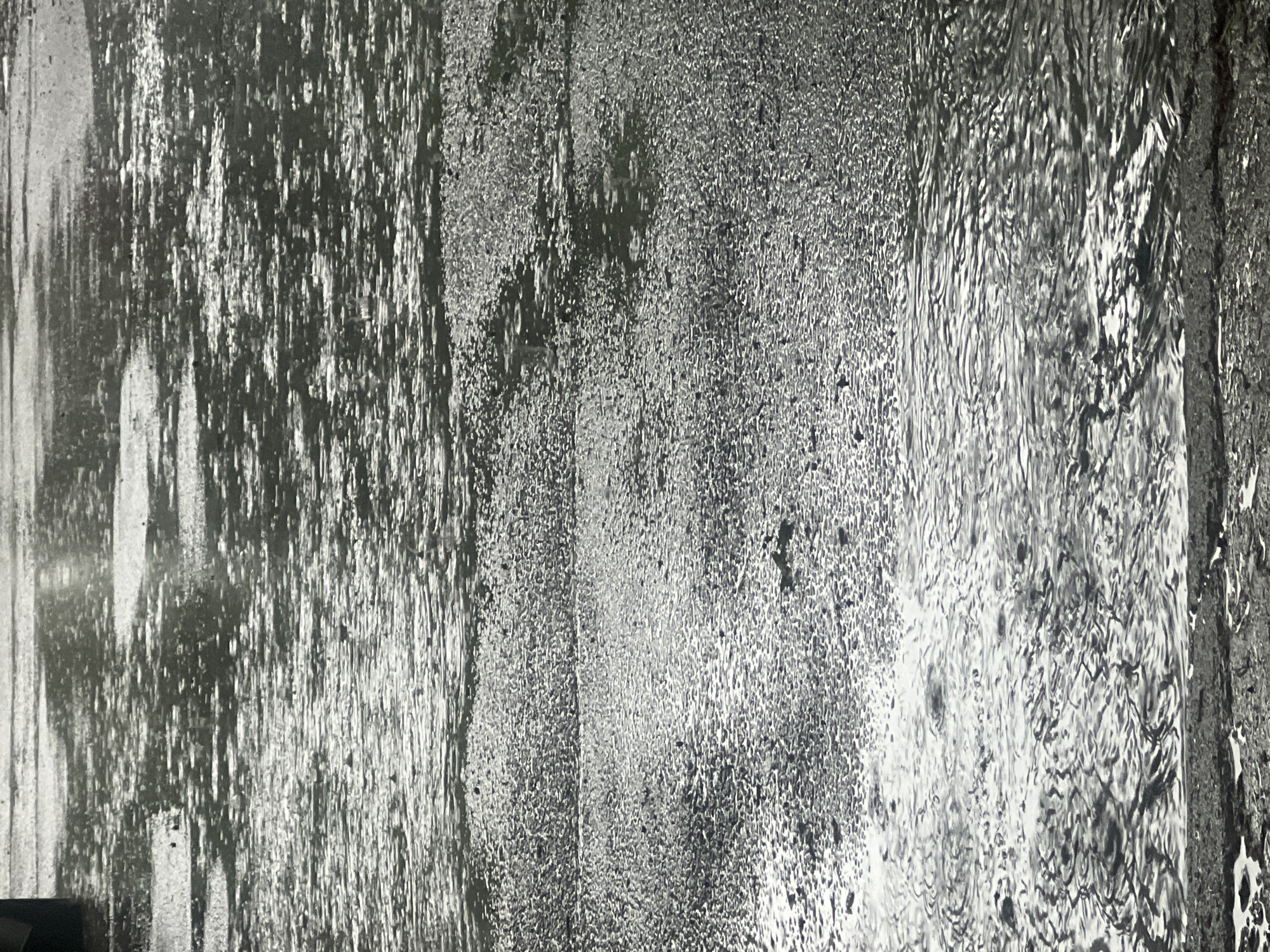 The image presents an abstract, grayscale close-up of a highly textured surface, suggestive of a wet or weathered environment. The composition is vertically oriented and divided into areas exhibiting distinct patterns.
On the left side, prominent vertical streaks and drips dominate, creating elongated, irregular formations of light and shadow, characteristic of water flowing down a surface. This transitions towards the center into a more granular, speckled texture, resembling condensation, frost, or a rough, aged material, with a dense scattering of small, bright points against darker tones. The rightmost section displays dynamic wavy and rippling patterns, strongly indicative of moving water or the intricate surface of a wet panel, like a waterfall or a water-streaked glass.
The entire image is rendered in shades of gray, black, and white, which accentuates the interplay of light and shadow on the varied textures. There are no discernible people, specific objects (other than the textured surface itself), or identifiable activities. The abstract nature provides no direct location cues within the image, though the context provided indicates it is from Sofia, Bulgaria. No visible text is present.