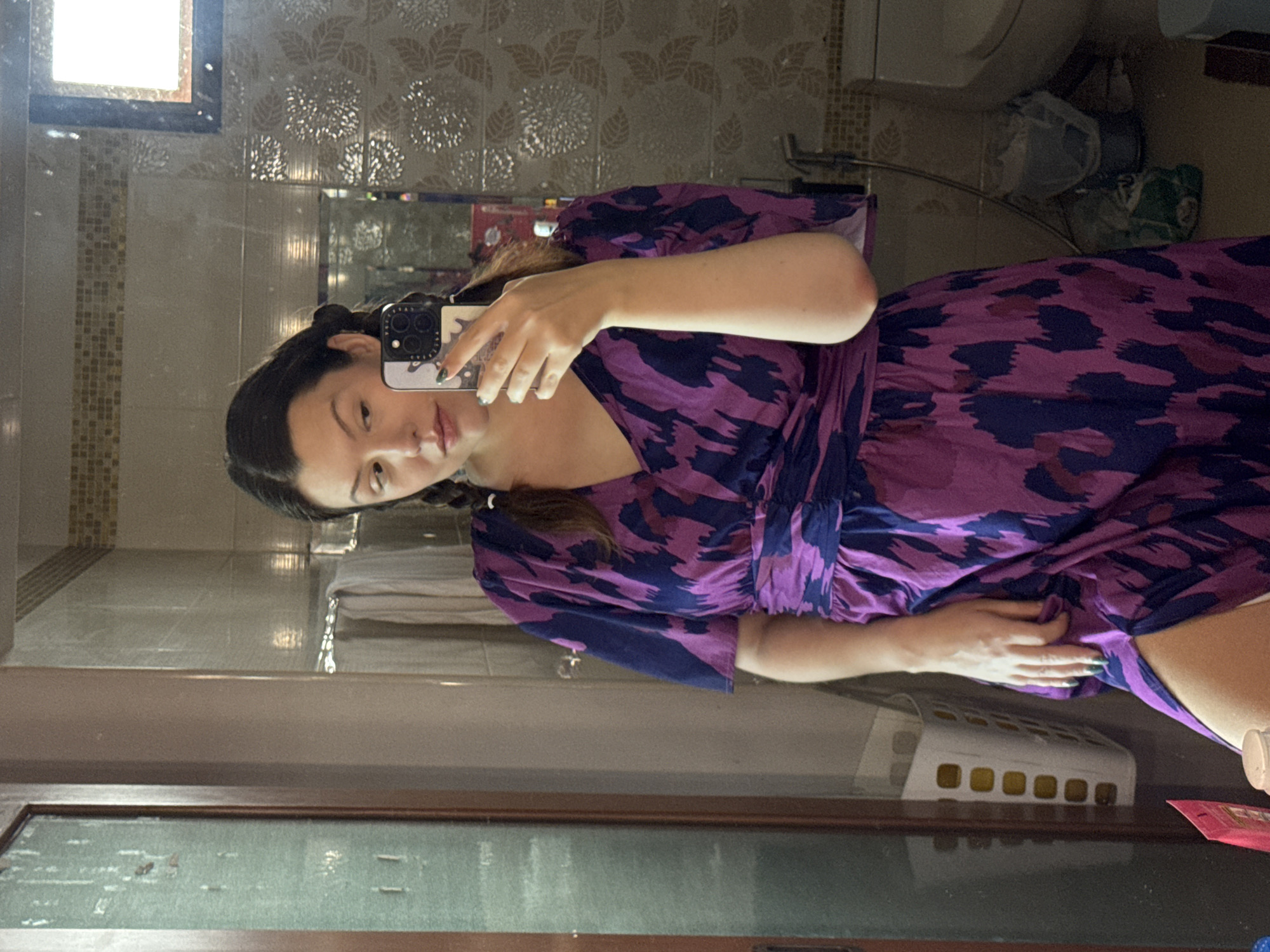 A woman is taking a mirror selfie in a bathroom. She is wearing a purple and blue patterned dress and has her hair in two braids. She is holding a phone in her right hand, and her left hand is resting on her hip. The bathroom appears to be small and dimly lit, with tiled walls and a toilet visible in the background. There is also a laundry basket and some cleaning supplies in the scene. The location is identified as San Phra Net Subdistrict, Thailand, based on the provided context, although this is not visually evident from the image itself.