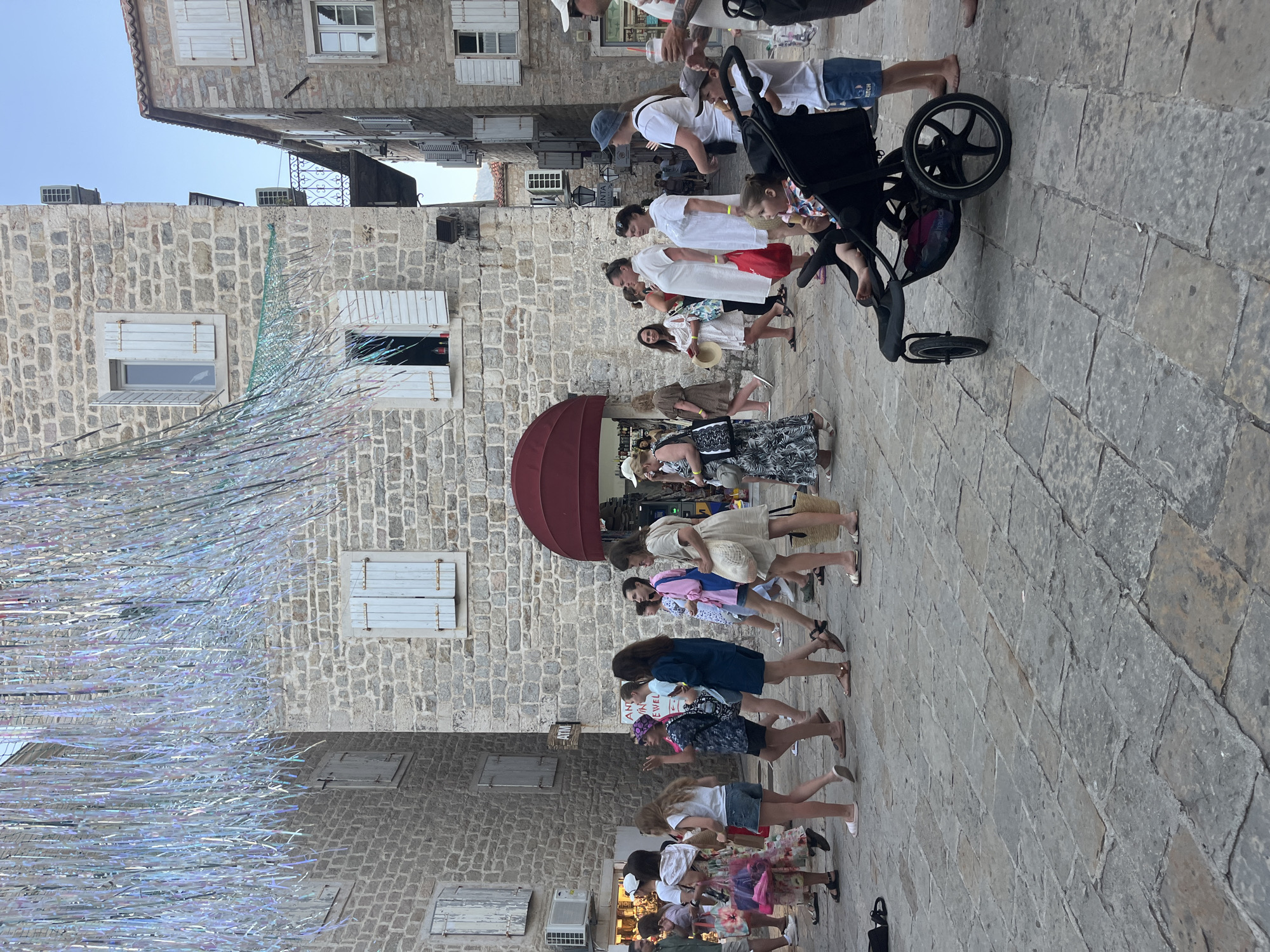 The image depicts a street scene in Budva, Montenegro, characterized by old stone buildings with light-colored shutters and a cobblestone pavement. A group of people, including women and children, are walking and standing in the street. Some of the individuals are carrying bags, and one woman is pushing a stroller with a child inside. Decorative, shimmering streamers are suspended overhead, suggesting a festive occasion or event. The architecture indicates a historic European setting, consistent with the Old Town of Budva. The overall atmosphere appears to be relaxed and casual, with people enjoying an outdoor environment.