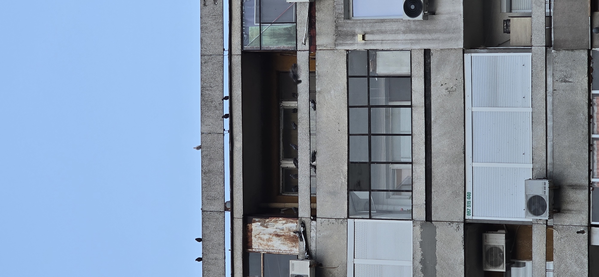 A building facade in Burgas, Bulgaria. The facade has a brutalist design with concrete sections. Windows are positioned between the sections. Pigeons are on the facade, especially in the open sections of the building. Air conditioning units are attached to the facade, with a phone number on one of them. The sky is blue.