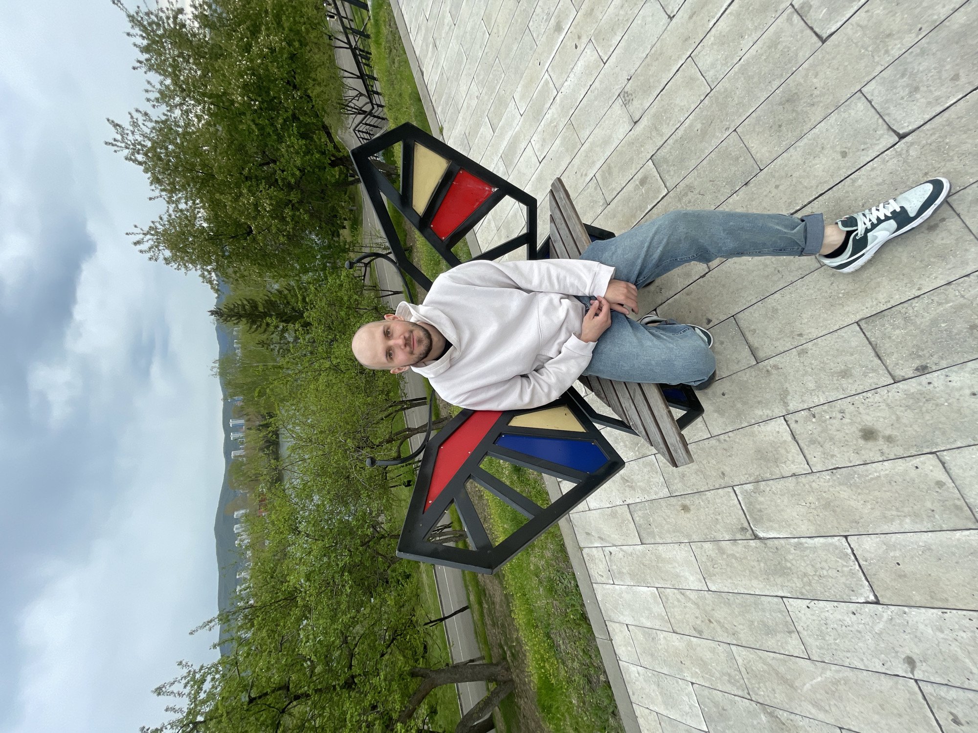 A man sits on a bench in a park, likely in Krasnoyarsk, Russia. He has a shaved head, a light beard, and is wearing a white hoodie, blue jeans, and green and white sneakers. The bench has a unique design with triangular colored inserts (red, blue, yellow) in the backrest. The park has trees with green foliage. The ground is paved with light grey rectangular stones. In the background, a city skyline is visible under a cloudy sky.