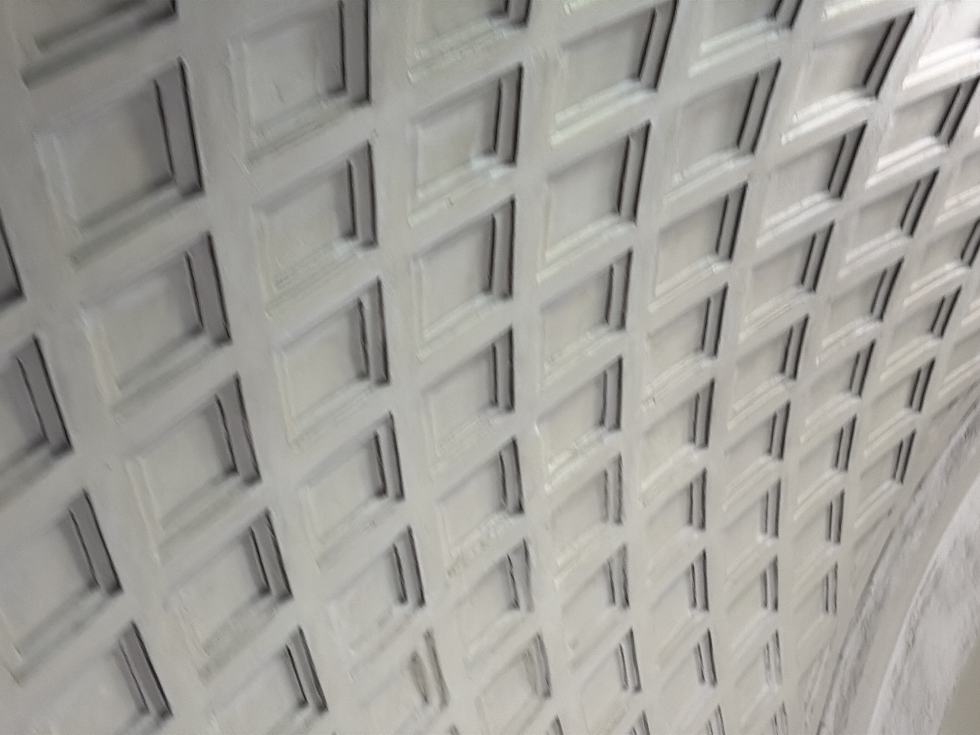 An angled, low-resolution photograph shows the white, coffered ceiling of a tunnel or underpass. The ceiling features a repeating pattern of rectangular indentations with beveled edges, casting subtle shadows that create depth. The perspective suggests the viewer is looking upwards and slightly away from the center of the tunnel. The overall impression is one of clean, repeating architectural elements, possibly a subway station or pedestrian walkway. The location is identified as Unknown, Unknown.