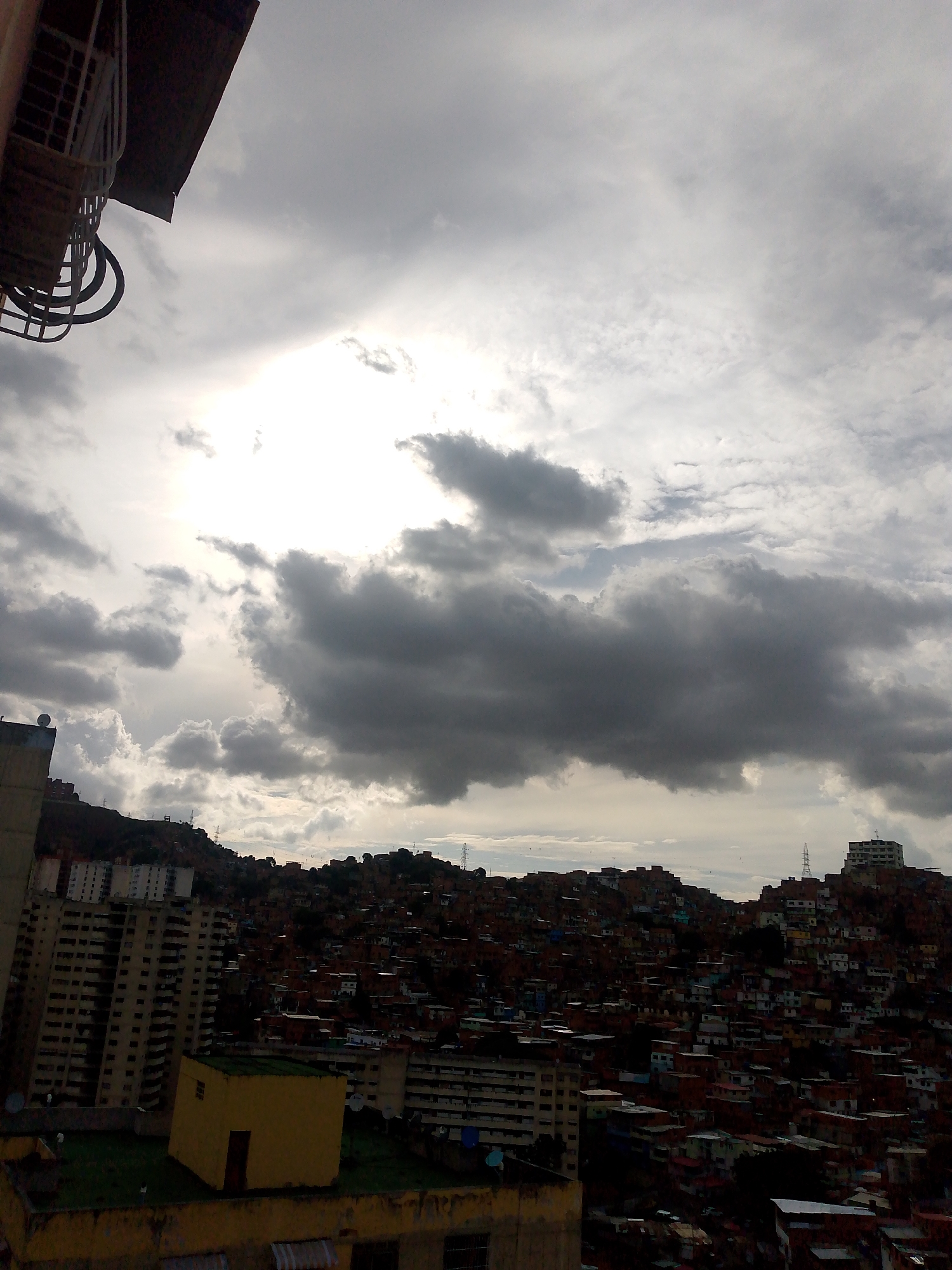 The image shows an overcast view of a densely populated hillside in Caracas, Venezuela. Many small buildings and houses, primarily red-brick or concrete, cover the slope. Larger apartment buildings are visible in the foreground and midground. The sky is filled with heavy, dark clouds obscuring the sun. A section of a building with metal railings and part of the building’s roof is in the upper left corner. The overall impression is one of urban density and heavy weather.