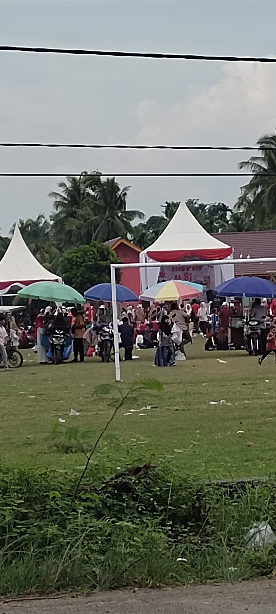 The image depicts an outdoor gathering in Peurupok, Indonesia. The scene features a grassy field with several tents, some white and red, others with umbrellas of various colors providing shade. A crowd of people, dressed in casual attire, are milling around. Some individuals appear to be standing or sitting, while others are in motion. Several motorcycles are parked in the area, indicating transportation. In the background, palm trees and other vegetation are visible, suggesting a tropical or subtropical environment. The sky is overcast, with some clouds present. The overall atmosphere suggests a community event or festival.