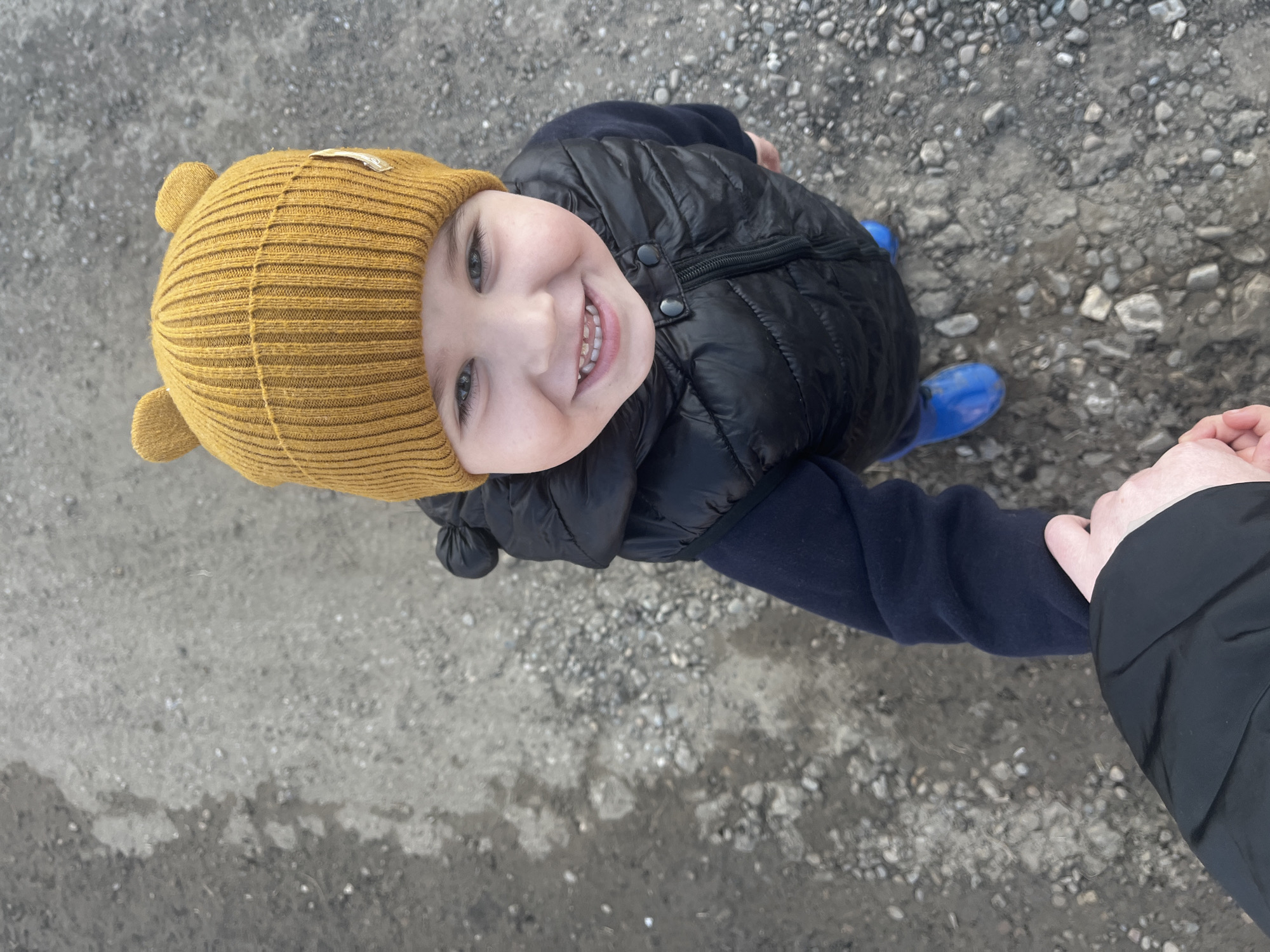 A high-angle shot depicts a young child holding someone's hand. The child wears a yellow knit cap with ears, a navy blue sweater, a black puffy vest, and blue rain boots. The child smiles, revealing their teeth. The setting appears to be an outdoor area covered in gravel and dirt. The context of Павловка, Russia is not evident from the image.
