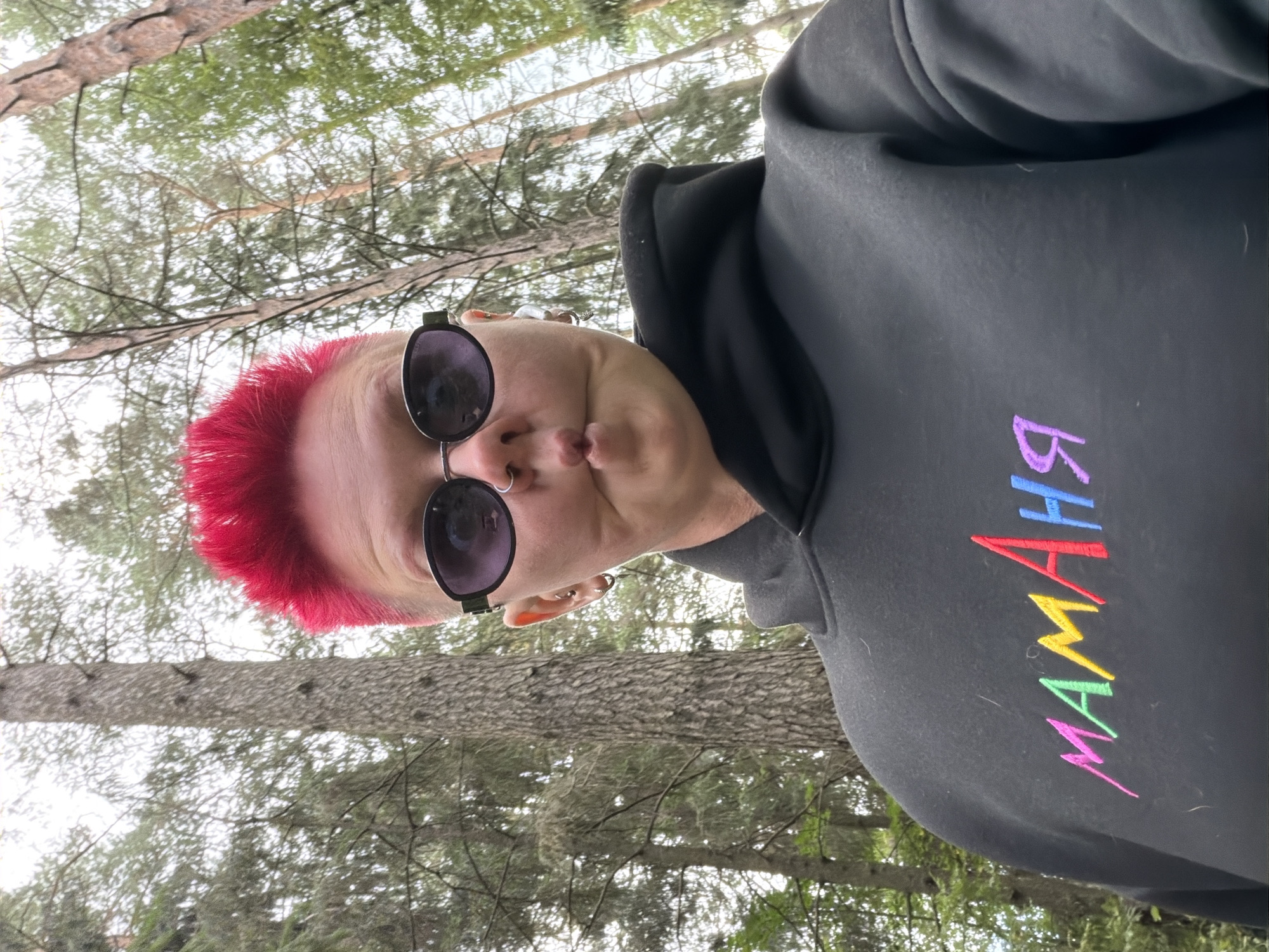 A woman with bright pink, spiked hair and sunglasses poses for a selfie outdoors, with a backdrop of trees and a cloudy sky. She is wearing a black hoodie with the word "МАМАНЯ" embroidered in colorful letters across the chest. Her facial expression is playful, with pursed lips. The setting appears to be a wooded area. The image does not provide any specific visual cues to determine its location within the city of Chaikovsky, Russia.