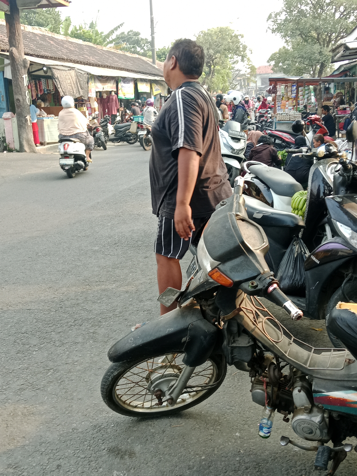 The image depicts a street scene in Kediri City, Indonesia. In the foreground, a man in a dark t-shirt and striped shorts stands beside a parked motorcycle. The motorcycle has a visible engine, a black fender, and a silver wheel. Behind him, the street is lined with shops or stalls, some displaying textiles and other goods. Several other motorcycles are parked or in motion on the street, carrying passengers. A woman in a light-colored top and patterned pants rides a white scooter in the middle ground, and other individuals are visible in the background, some on motorcycles and others near the stalls. The environment suggests a bustling marketplace or commercial area with typical street traffic. The overall lighting indicates it is daytime.