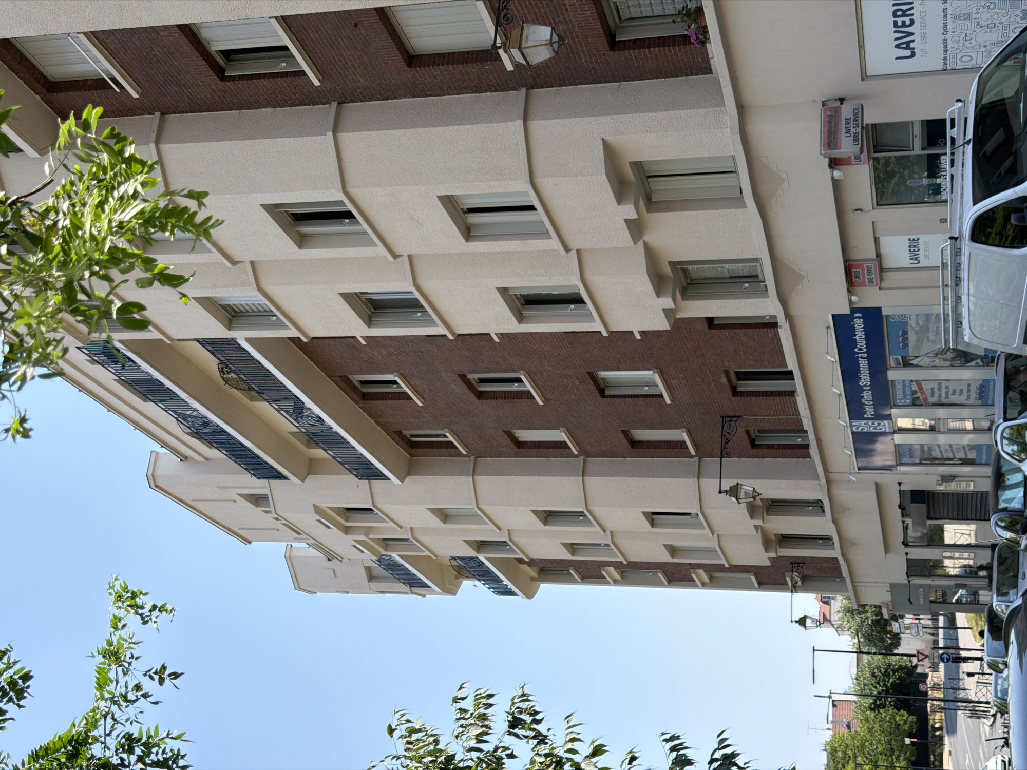 This is a street-level photograph, taken from a low angle looking upwards, of a multi-story building. The building has a facade of alternating sections of reddish-brown brick and cream-colored stucco. The stucco sections feature projecting horizontal bands of varying depths, creating a layered effect. There are numerous windows visible across the facade, some with dark frames and others appearing to be recessed. Small balconies with dark railings are present on some levels. In the foreground, on the left side of the image, is a cluster of green leafy branches, suggesting that the photograph was taken during a time of year when trees are in full foliage. The sky above is a clear, bright blue, indicating daytime.
On the ground floor of the building, there are several commercial establishments. One prominent sign reads "LAVERIE LIBRE-SERVICE" in large letters, indicating a self-service laundry. Other signs are partially visible, including "MACHINES A LAVER" (washing machines) and what appears to be a "Point d'Info Stationner à Courbevoie" (Information point Parking in Courbevoie), suggesting the location is Courbevoie, France, and possibly related to parking information or services. A white commercial van is parked on the right side of the street, partially obscuring the ground-level storefronts. Streetlights and traffic signs are visible further down the street, hinting at an urban environment. The overall impression is a mundane urban street scene on a bright, clear day.