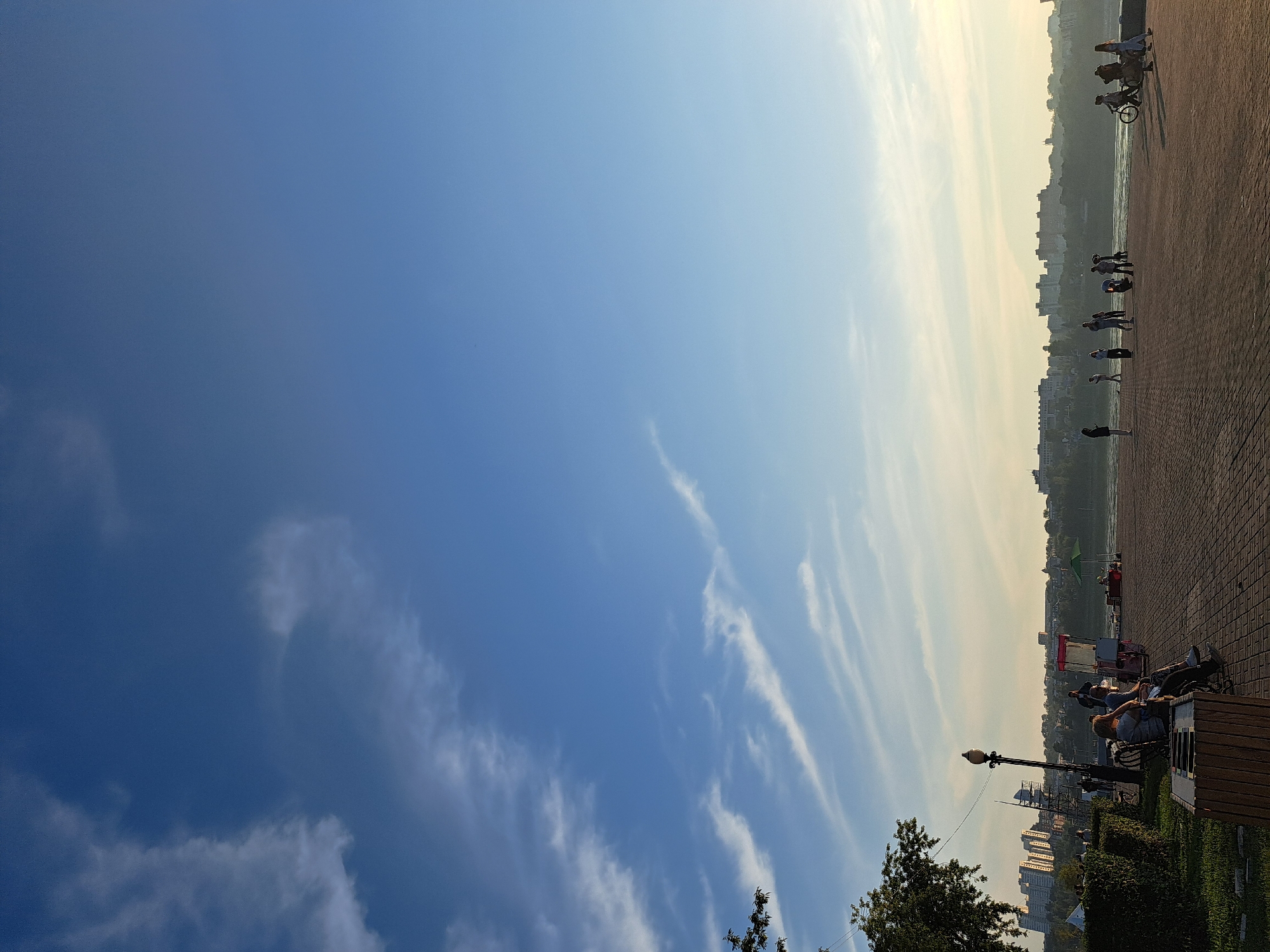 The image depicts a scene on a paved walkway next to a body of water, likely a river or lake, in Irkutsk, Russia. In the background, a city skyline with numerous buildings is visible across the water, partially obscured by trees. The sky is blue with scattered wispy clouds, illuminated by the warm light of either sunrise or sunset.
On the walkway, several people are scattered. In the foreground, a few individuals are seated on a bench. Further along the walkway, groups of people are standing and walking. One person is standing next to a bicycle, and another appears to be pushing a cart. There are also some vendor stalls or kiosks visible, one with a red and white structure. Green bushes and trees line the left side of the walkway, and a lamppost stands near the bench. The walkway itself is paved with interlocking bricks. The overall impression is of a public space being utilized by people during a pleasant time of day.