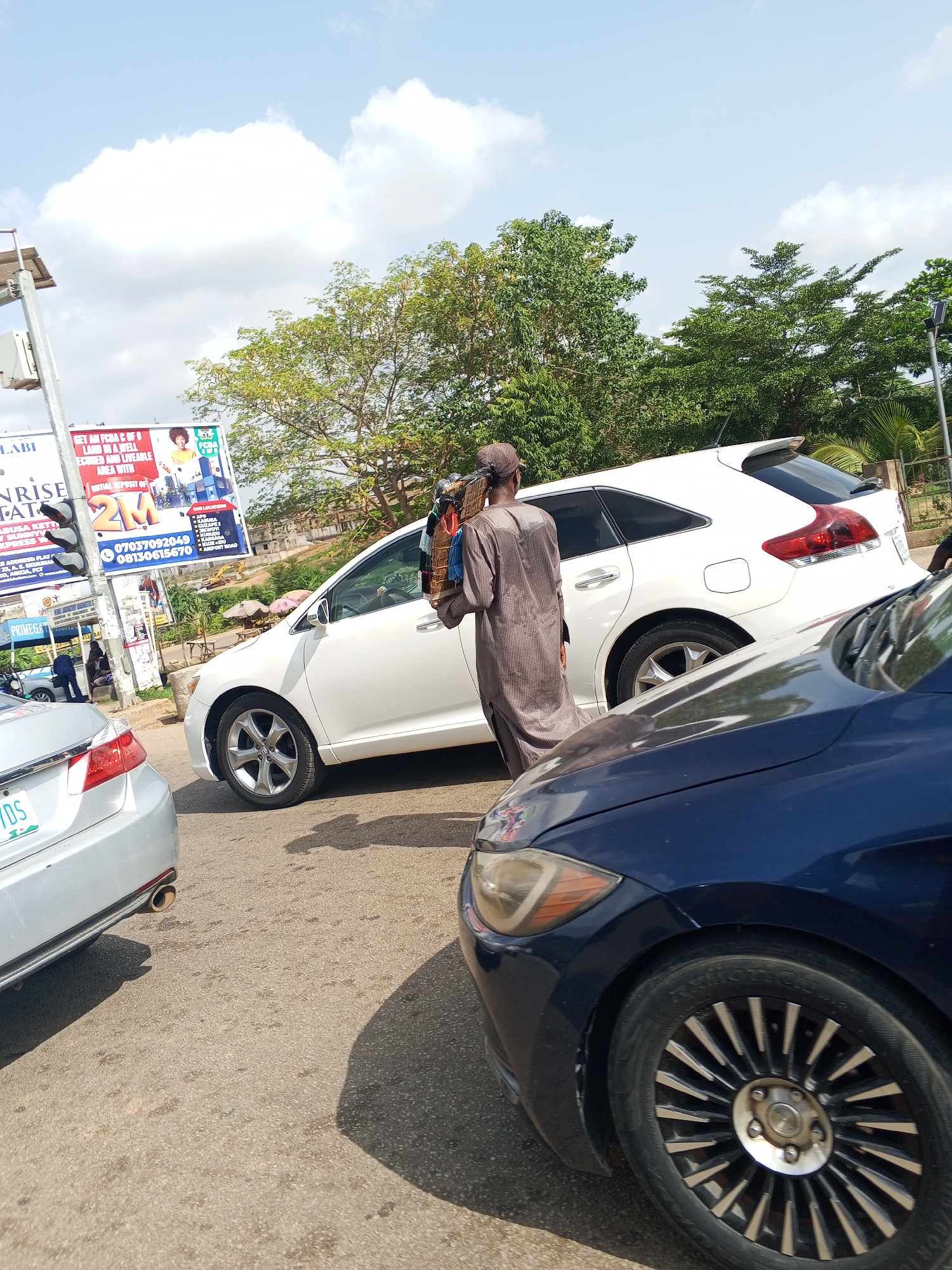 The image captures a street scene in Abuja, Nigeria, with a man walking between cars in a traffic jam. The man is wearing a brown traditional robe and a hat, carrying a loaded rack on his back. Two cars are visible: a silver sedan with a green license plate ("17DS") and a blue sedan. A white SUV is in the background. Billboards advertising real estate in Abuja are visible in the background. The text on the billboards includes contact information for real estate agencies. The sky is bright with scattered clouds, and there are trees alongside the road.