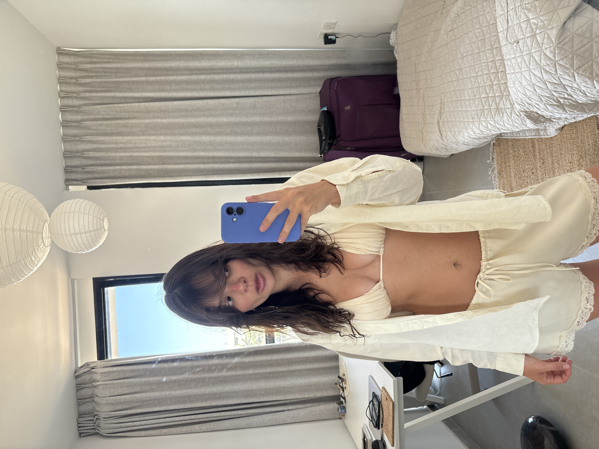 A young woman is depicted taking a mirror selfie inside a brightly lit room, stated to be located in Tel-Aviv, Israel. She has dark, wavy hair with bangs and is holding a blue smartphone with a visible dual-lens camera in her right hand. Her expression is characterized by a slightly open mouth as she looks towards the mirror. She wears a cream-colored string bikini top, an unbuttoned, loose-fitting cream long-sleeved shirt, and matching cream shorts with a drawstring waist and lace trim at the hem. A subtle nose ring is visible.
The room features plain white walls and a light gray tiled floor. On the left side, a large window with a black frame allows ample natural light into the room, revealing a bright blue sky and distant modern architecture outside; light grey pleated curtains partially obscure the view. Two white, round paper lantern-style light fixtures hang from the ceiling.
To the left, a white modern desk holds items including sunglasses, a cork coaster, and a dark cylindrical object. A grey office chair is partially visible beneath the desk. To the right, a single bed covered with a light beige quilted bedspread is positioned against the wall. A small, textured woven rug lies on the floor next to the bed. Between the desk and the bed, a large purple suitcase with a black bag resting on top is situated against the wall. A white electrical outlet with a black charger plugged in is visible on the wall above the bed.