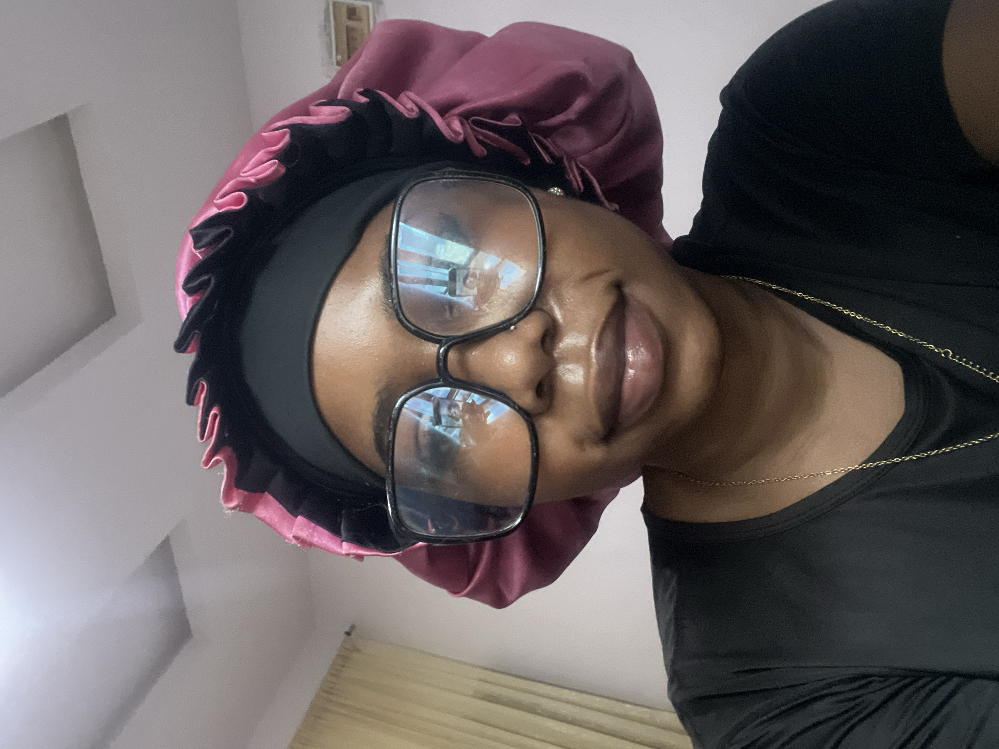 The image depicts an indoor, eye-level shot of an individual in Rafin Par, Nigeria. The subject, a person with dark skin, is centered, wearing a black short-sleeved t-shirt and a thin gold chain necklace. A black headwrap is visible beneath a voluminous, ruffled pink satin bonnet. Large, black-framed rectangular glasses cover the subject's eyes, exhibiting significant reflections. Within the reflections in both lenses, an overhead rectangular ceiling light fixture is prominent. The right lens also shows a partial reflection of a person holding a smartphone, suggesting the image is a selfie. The subject displays a subtle smile with glossy lips. The background consists of a plain, light-colored wall. To the left, beige curtains are partially visible. The ceiling is white, featuring a rectangular recessed area. A small, white rectangular object, possibly a light switch or outlet, is affixed to the wall on the far right.