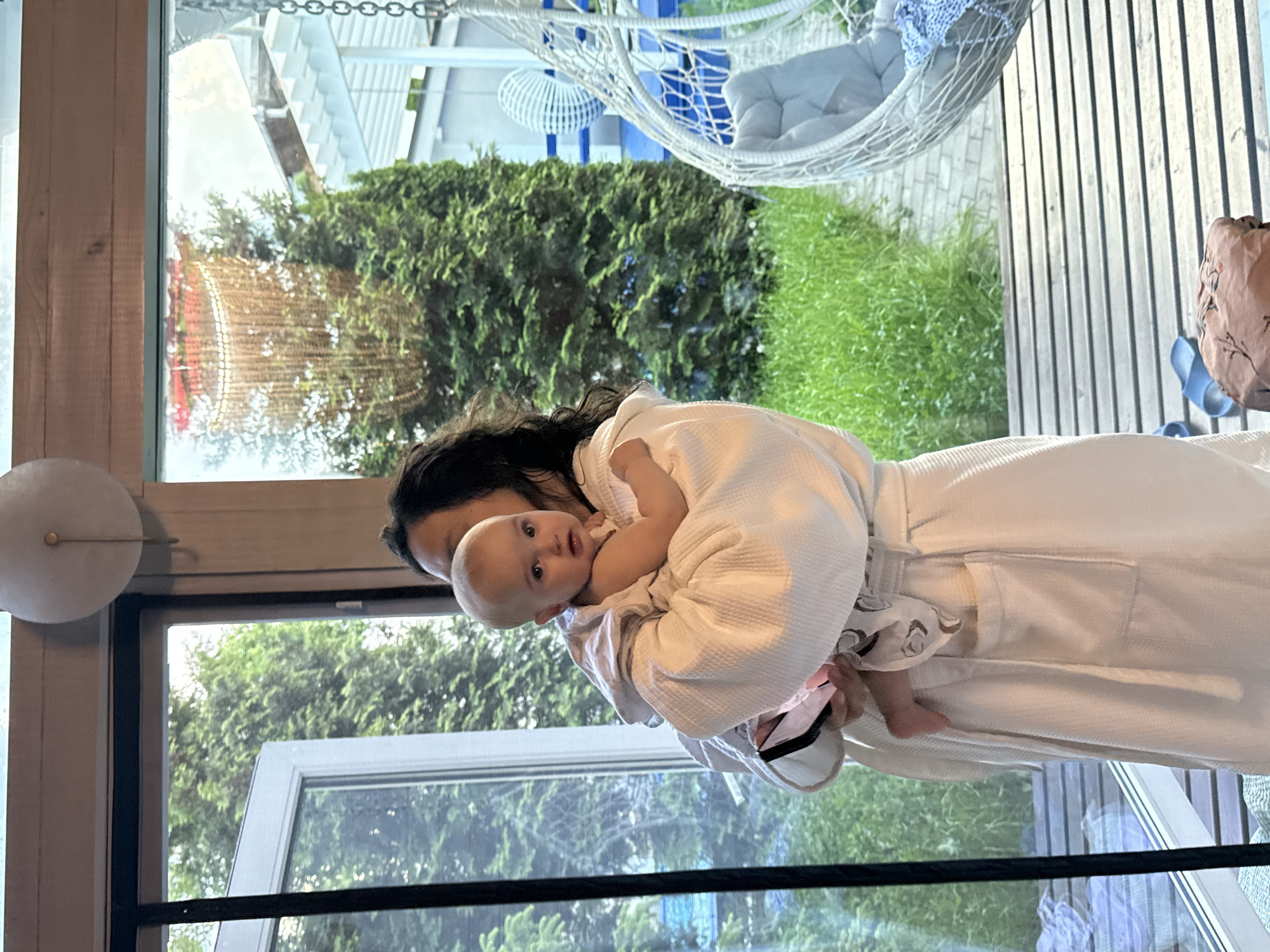 A person in a white robe is holding a baby, likely indoors. Outside, a garden is visible through a window, including grass, trees, and a white wicker chair. Based on the location Чёрное, Russia, and the reflection in the window, the building with red accents and lights could be a restaurant or establishment named "The Black Pearl."