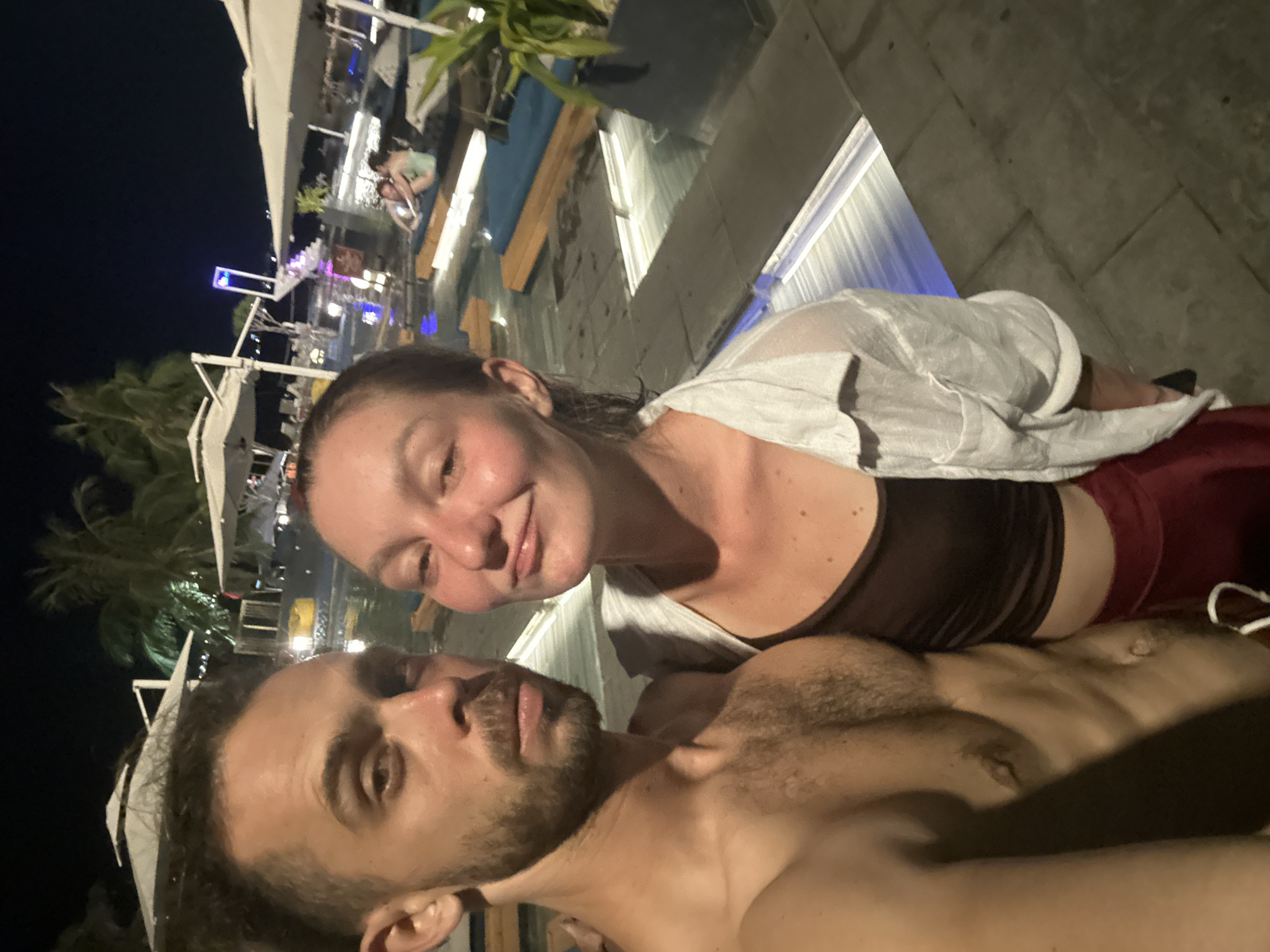 This is a selfie taken at night, possibly in Đà Nẵng, Vietnam, given the context. The image features a man and a woman posing closely together. The man, in the foreground, has a beard and is shirtless. The woman is behind him, wearing a white sheer top over a dark colored camisole and a red skirt. Her face is turned towards the camera, and she has a slight smile.
The background suggests an outdoor evening setting, possibly a resort or a restaurant area with a pool. There are lights illuminating the area, including some blue and white lights around what appears to be a pool or water feature. Umbrellas and seating areas are visible in the distance, indicating a place for relaxation or dining. Palm trees can also be seen, consistent with a tropical or subtropical location like Đà Nẵng.
The lighting is dim, as expected for nighttime, with artificial lights providing illumination. The overall mood appears relaxed and intimate, with the couple close together and smiling. There is no visible text in the image.