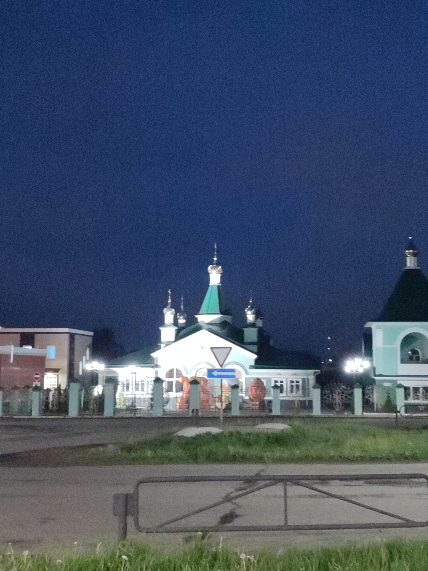 At night in an unknown city, an Orthodox church stands illuminated. The church features green roofs, domes, and bright white walls. A blue and white directional sign is visible in front. The area is surrounded by a fence with trees, and a road runs in front of the scene, obstructed by a metal barrier. A building with faint lighting is seen on the left.