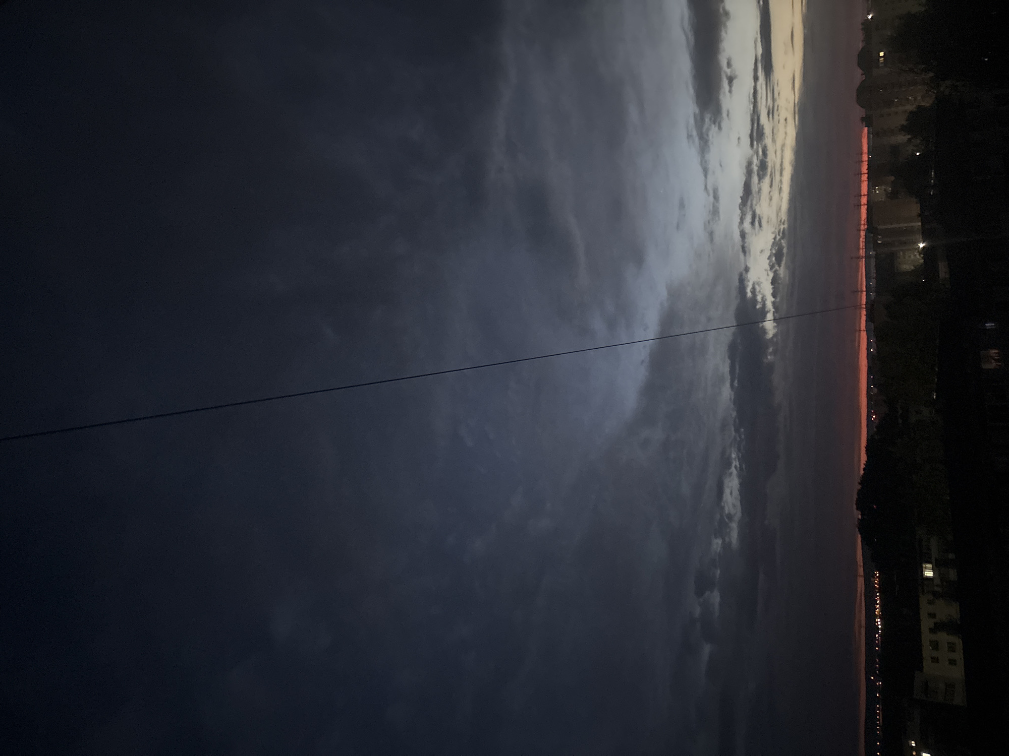 The image depicts a dark, evening or nighttime scene, likely in Chaikovsky, Russia. The sky is predominantly dark blue and filled with clouds. A wire crosses the image diagonally. On the right side, there's a faint orange glow on the horizon, possibly from the setting sun. Dark silhouettes of buildings and trees are visible against the skyline. Streetlights are scattered and can be seen illuminating buildings in the city.
