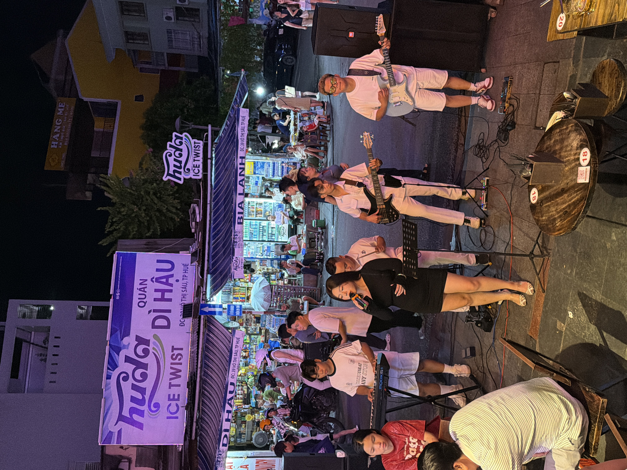 The image captures a nighttime scene on a street in Thuận Hóa Ward, Vietnam, likely a commercial area with shops and restaurants. In the foreground, a live band is performing. A female singer, dressed in a black mini-dress, is holding a microphone and singing. To her left, a young man is playing a keyboard, and to her right, another young man is playing a light blue electric guitar. Further to the right, a third young man is playing a dark electric bass guitar. All the musicians are wearing white shirts and pants. The band is set up on the street, with musical equipment including speakers, amplifiers, and cables visible.
Behind the band, a street-side shop or restaurant is open, with customers seated at tables and standing around. The shop has a striped awning and shelves stocked with various goods. A sign above the shop reads "QUÁN DÌ HẬU ICE TWIST" in large letters, with smaller text below indicating the address "ĐC: 39 Võ Thị Sáu, TP. Huế". This sign also features the logo for "huda" and "ICE TWIST". Another sign further up the street, illuminated in yellow, reads "HANG ME" and appears to advertise food specialties.
Several people, including customers and passersby, are visible in the background. A motorcycle is parked on the street, and other vehicles are present in the distance. The lighting is a mix of streetlights, shop lights, and illuminated signs, creating a vibrant, urban atmosphere. The ground is paved with concrete and asphalt. The overall scene depicts a lively evening in a Vietnamese city, with a focus on local businesses and street entertainment.