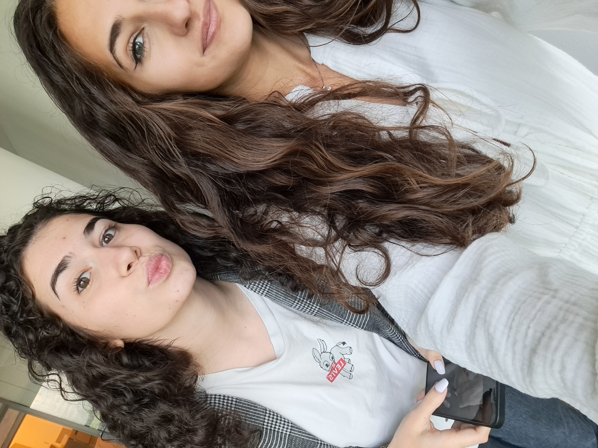 Two young women are posing for a selfie indoors. The woman on the bottom is holding a black smartphone, wearing a white t-shirt with a cartoon bunny, and a grey plaid jacket. Her hair is dark and curly, and she is making a pouting face. The woman on top has long, wavy brown hair, and is wearing a white shirt. She has light skin and light-colored eyes. The background appears to be a white wall or fabric. The location in Sofia, Bulgaria is not apparent from the image alone.