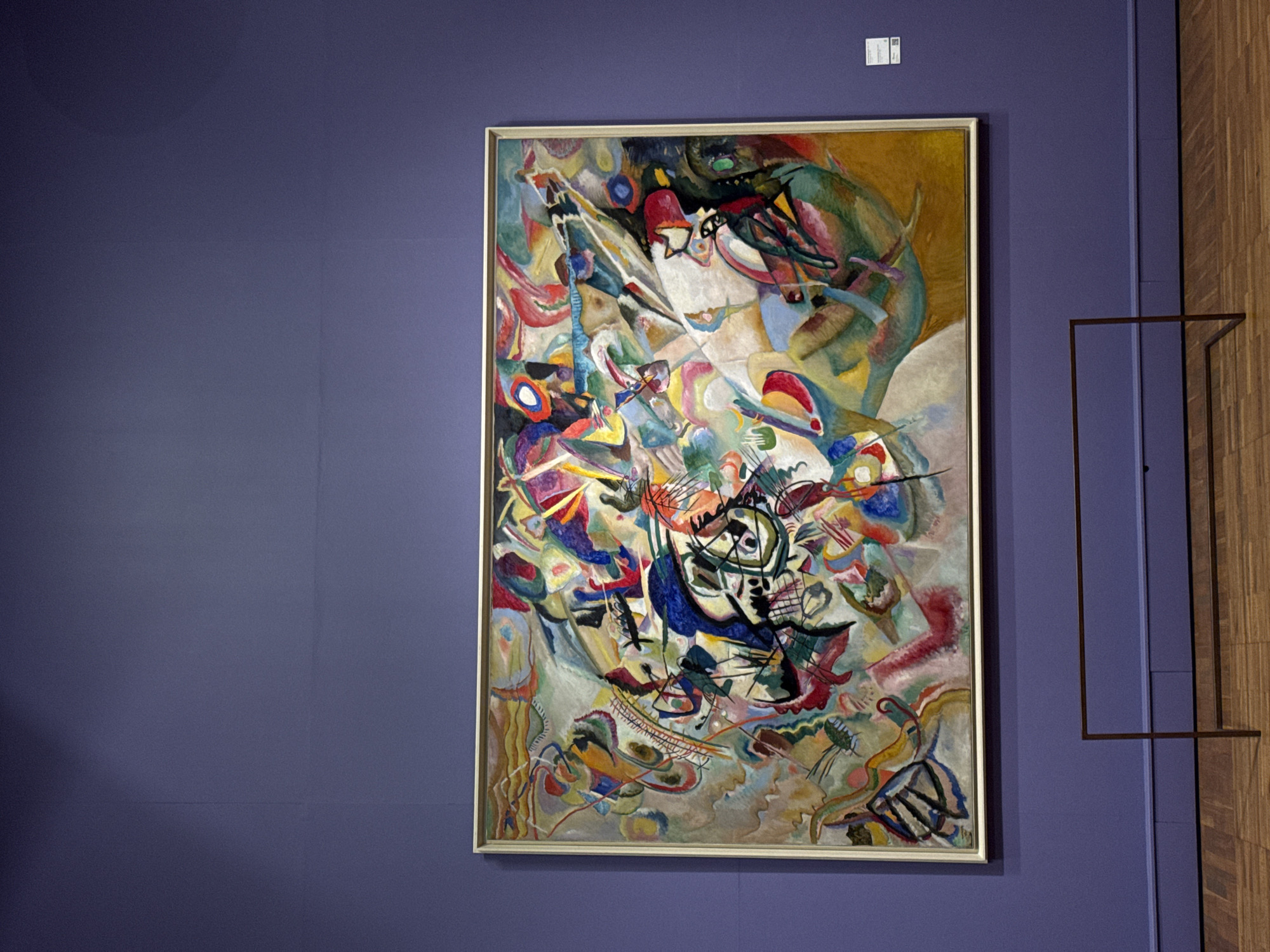 A painting hangs on a wall in a gallery. The painting is abstract and colorful, with many different shapes and colors. It is in a light gold frame. The wall is a deep blue-purple. To the right of the painting is a brown wooden wall with a dark brown minimalist rectangular wall mount. The painting is located in the Tretyakov Gallery in Moscow, Russia.