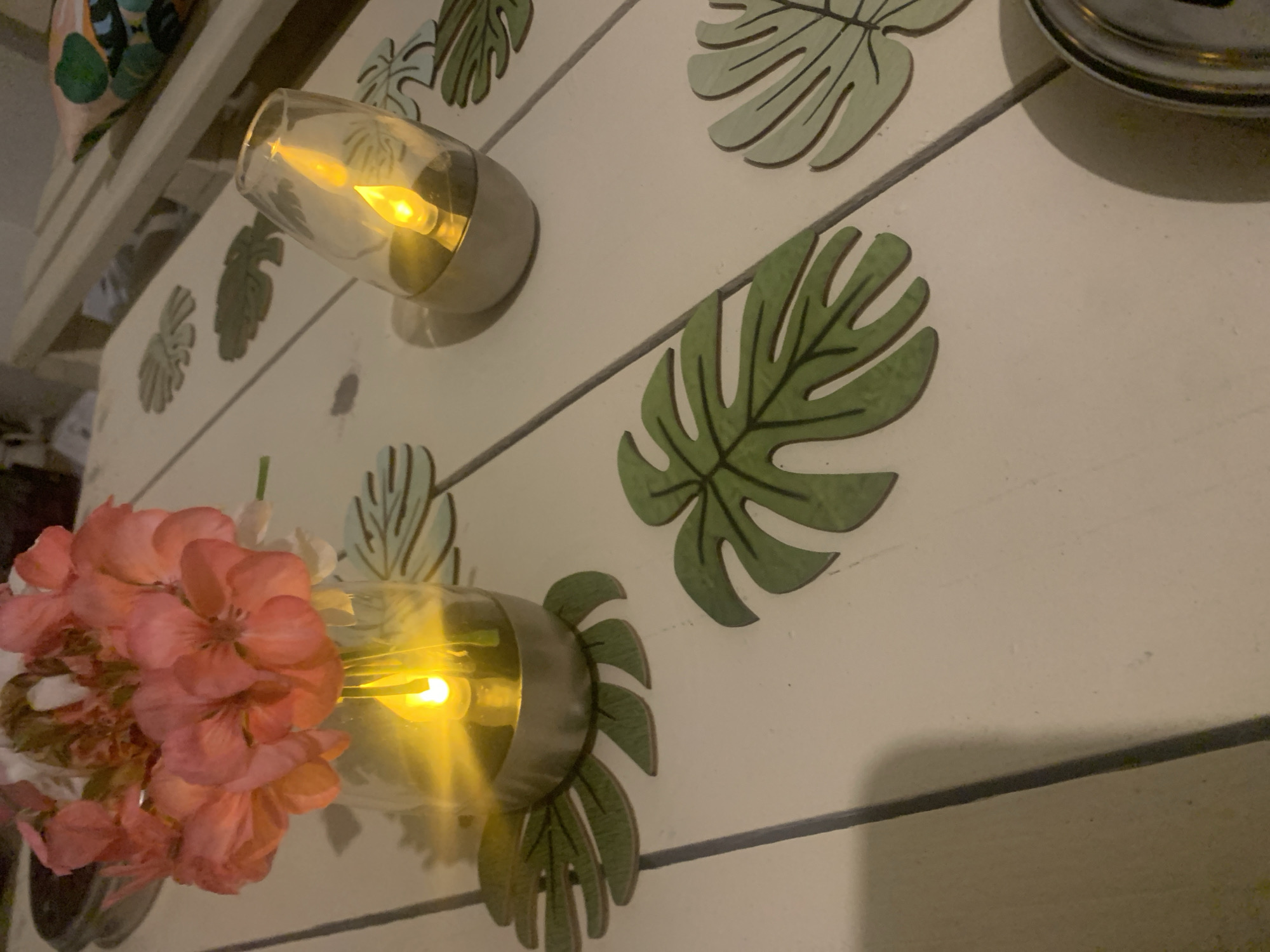 The image shows an outdoor table setting at night. The table is made of white planks and decorated with green monstera leaf coasters. On the table are two solar-powered LED lights designed to resemble flickering flames, one holding a bouquet of pink flowers. The scene appears to be a backyard or patio, possibly in the city of Khazar Raion, Azerbaijan, given the context. The lighting is dim, with the artificial flames casting a warm glow.