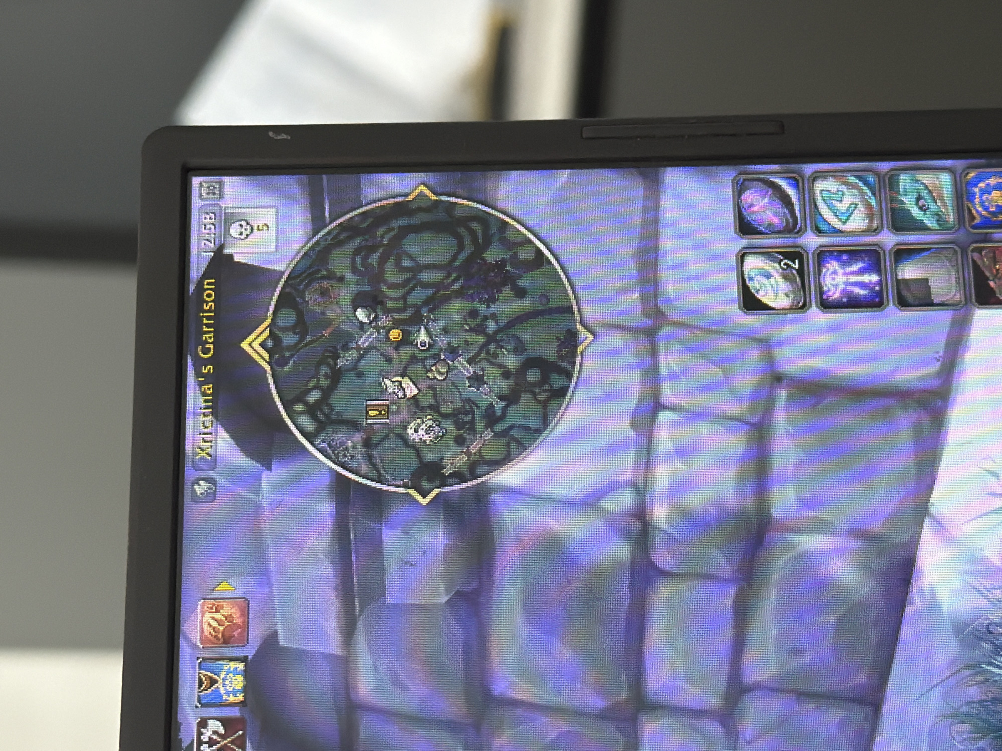 The image displays a close-up view of a computer screen, showing a video game interface. The game appears to be World of Warcraft, indicated by the presence of a minimap in the upper right quadrant of the screen, displaying a location labeled "Xristina's Garrison." The minimap features various icons representing in-game elements. Below the minimap, a set of action bars with game icons is visible on the right side of the screen. The background of the game interface shows a textured stone wall. The computer is positioned in an indoor environment, with a blurred background suggesting a room with natural light entering from the right. There are no people visible in the image. The screen exhibits a noticeable pixelation or grid pattern, characteristic of a digital display. The overall scene depicts a moment of gameplay captured on a screen. There is no information in the image to suggest any connection to Plovdiv, Bulgaria.