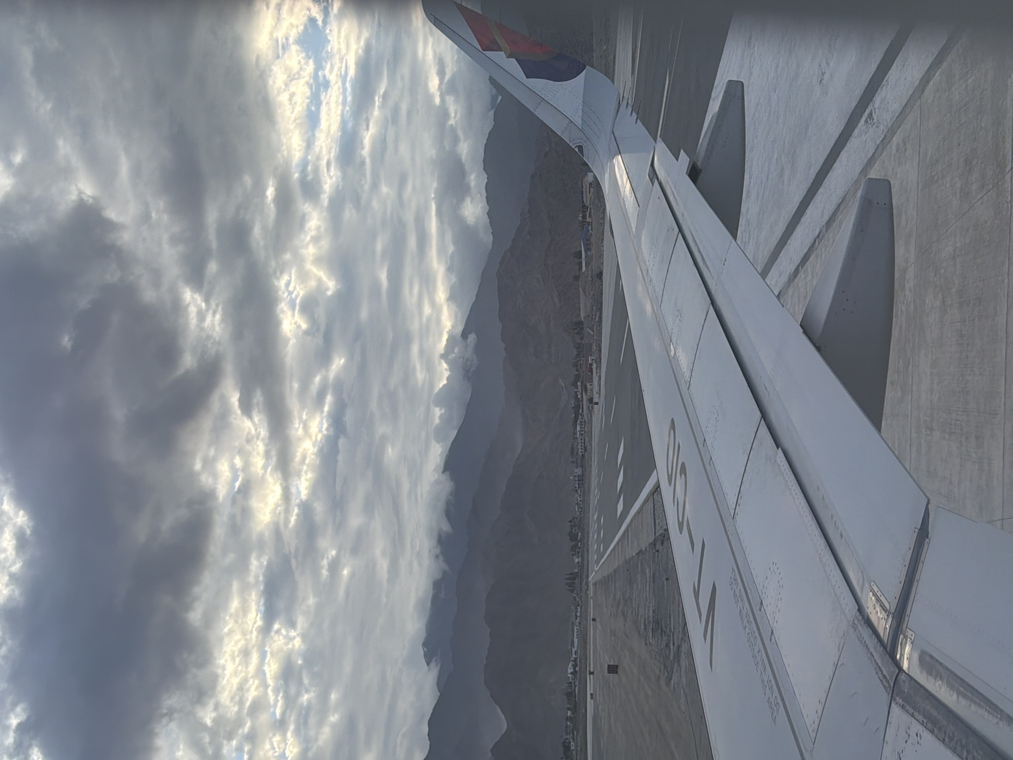 This is a photograph taken from the window of an airplane, likely a passenger jet, on a tarmac. The airplane's wing is prominently in the foreground, extending across the lower half of the image. The wing is white with some markings, including the letters "VT-CJO" visible on its surface. The wing appears to be attached to the fuselage, with visible rivets and panel lines.
The airplane is on a runway or taxiway at an airport. The tarmac is grey concrete with white markings indicating lanes and boundaries. In the middle ground, the runway stretches into the distance, lined with lights or markers. Beyond the runway, a desolate landscape unfolds, characterized by barren, mountainous terrain. The mountains are muted in color, appearing grey and brown, and are partially obscured by soft, white clouds that cling to their peaks. The sky above is overcast with a dramatic display of clouds. Dark grey clouds dominate the upper portion of the image, while lighter, wispy clouds with patches of sunlight breaking through are visible closer to the horizon, creating a sense of depth and atmospheric interest.
The scene suggests the airplane is either preparing for takeoff or has just landed. The lighting indicates it might be early morning or late afternoon, given the soft, diffused light and the overcast conditions. The location appears to be Spituk, India, which is known for its airport situated in a valley surrounded by the Himalayas. The barren, rugged mountains and the vast, cloudy sky are characteristic of the high-altitude desert environment of Ladakh. There are no people visible in the image, and the focus is on the airplane, the runway, and the surrounding natural landscape. The overall mood is serene and somewhat dramatic due to the expansive cloudy sky and the imposing mountains.