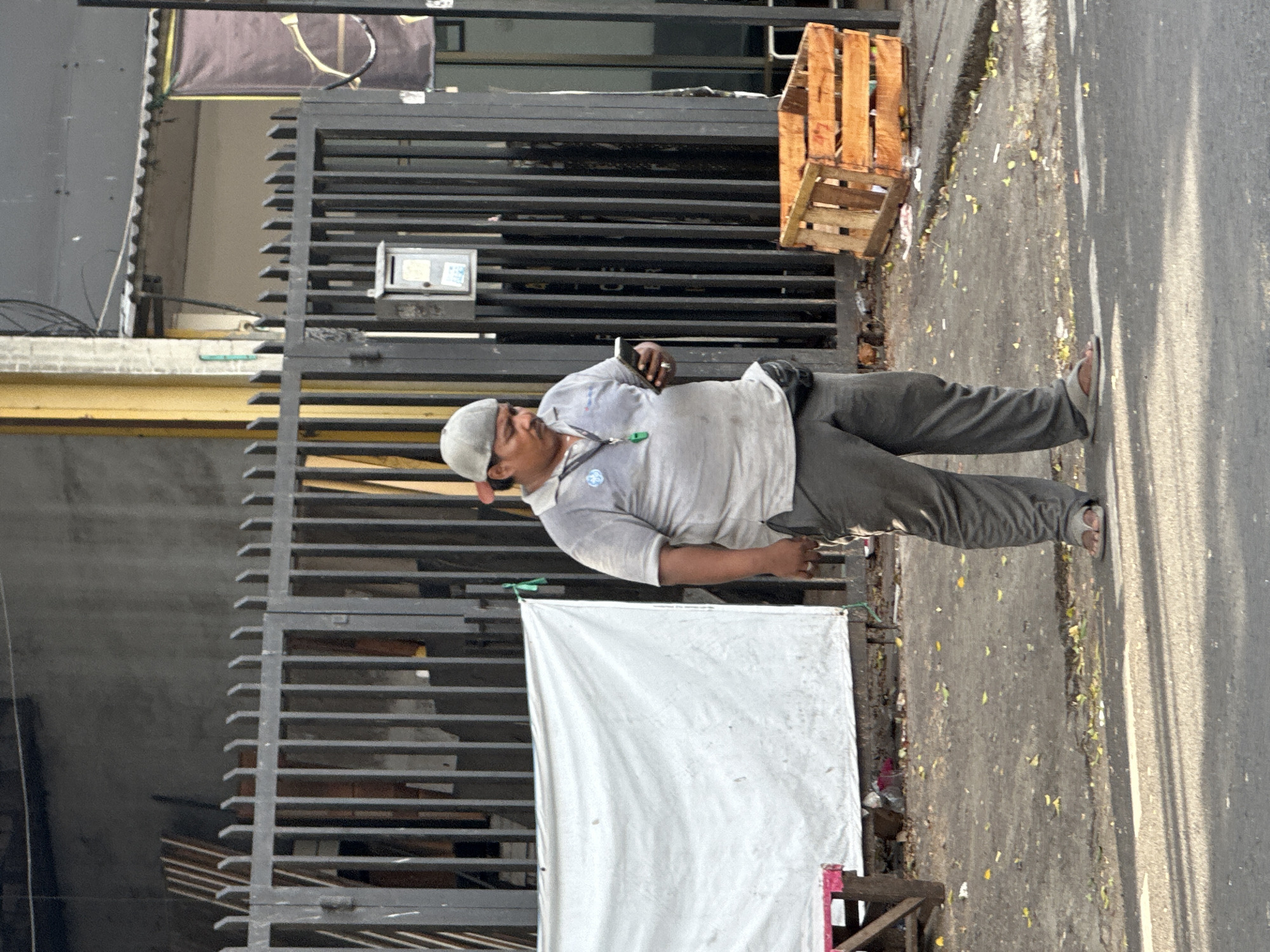 The image shows a man standing in Central Jakarta, Indonesia, next to a fence with a white cloth hanging on it. He is wearing a light gray shirt, dark gray pants, a gray cap, and sandals. He holds a mobile phone in his left hand. A wooden crate sits on the sidewalk beside the fence. The area appears to be a street with leaves scattered on the asphalt.