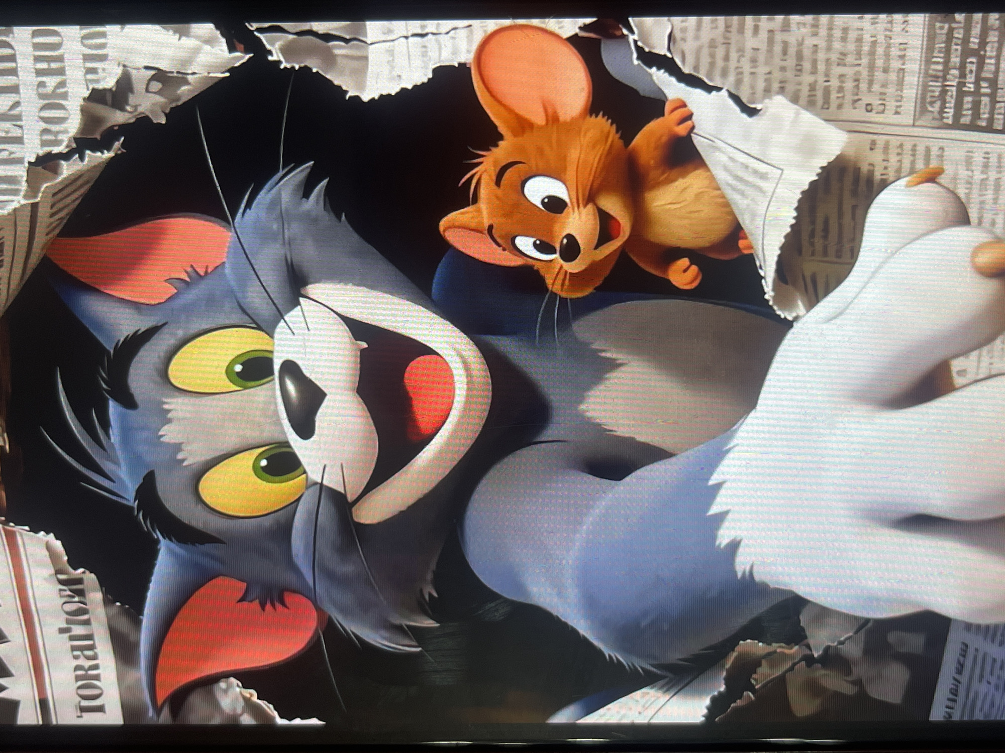 The image depicts a scene from the movie "Tom and Jerry" where the characters are breaking through a newspaper. Tom, a large blue cat, is on the left, and Jerry, a small brown mouse, is on the right. Both characters are smiling and appear to be engaged in a playful chase or escape. The background is a torn newspaper with visible text. There is no indication of Bishkek City, Kyrgyzstan, or any specific location within it.