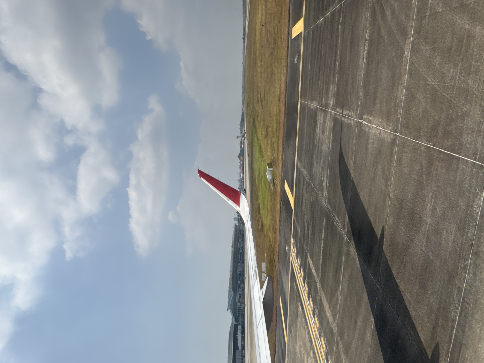 A view from an airplane window shows the wing of the plane with a red and white tail fin over an airport runway. The runway is marked with yellow lines. Grass borders the runway. In the distance are buildings resembling an airport terminal. The sky is partly cloudy. While it is difficult to pinpoint the exact location from the image, the query specifies Đà Nẵng, Vietnam, suggesting the image was taken at Đà Nẵng International Airport.