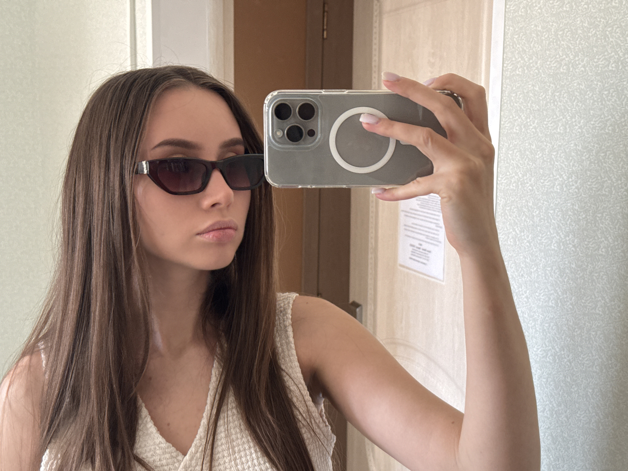 A young woman with long brown hair is taking a selfie with a phone in a light-colored room. She is wearing dark sunglasses and a white top. The background consists of a door and patterned wall. The location appears to be indoors, possibly a room in Береговой сельский совет, Ukraine, although the exact address cannot be determined from the image.