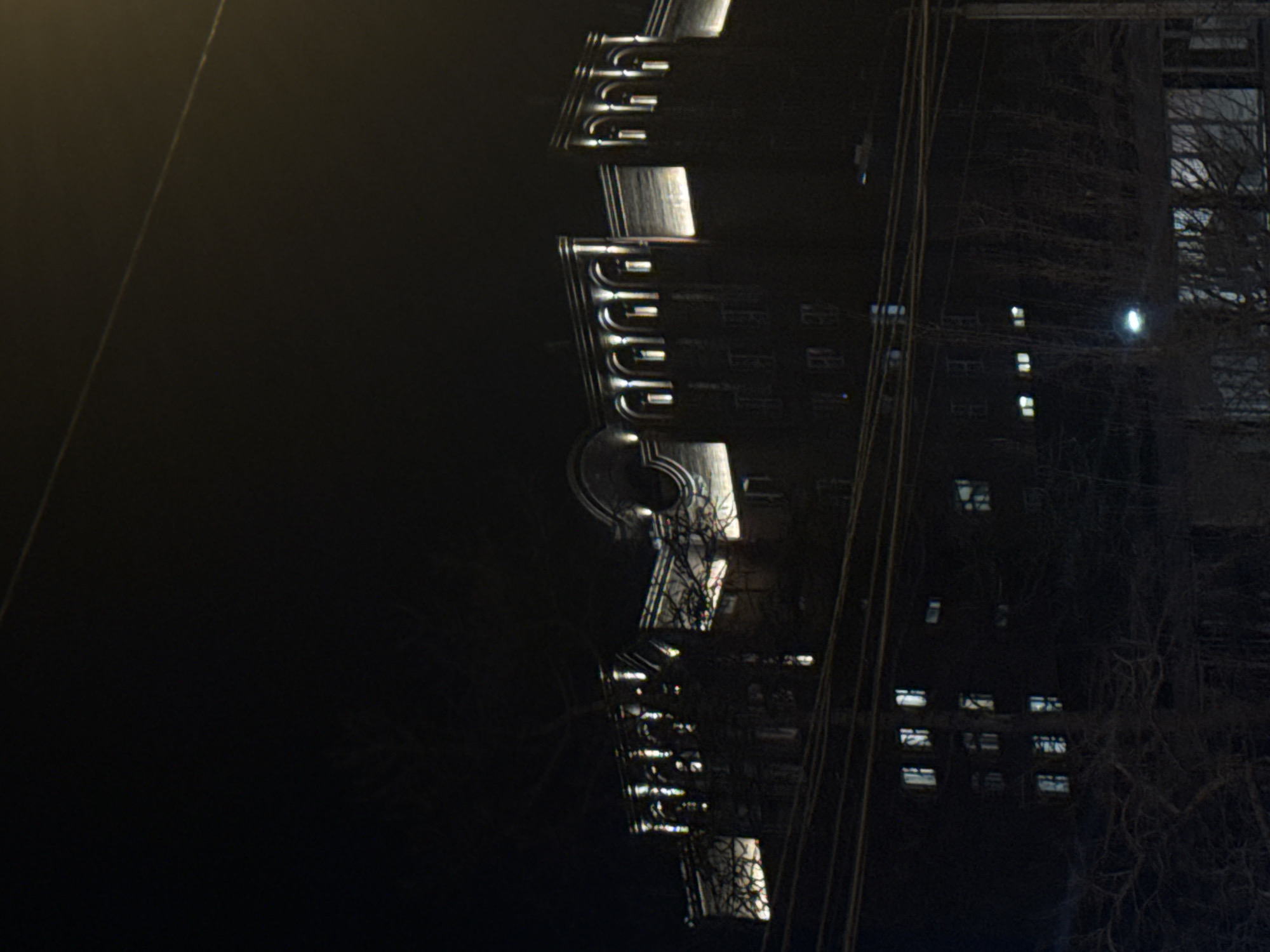 The image shows a tall building in Krasnoyarsk, Russia, at night. The building's facade is illuminated, highlighting architectural details. There are also rows of lit windows. Power lines are visible in the foreground, and bare tree branches partially obscure the building. The sky is dark, suggesting nighttime.