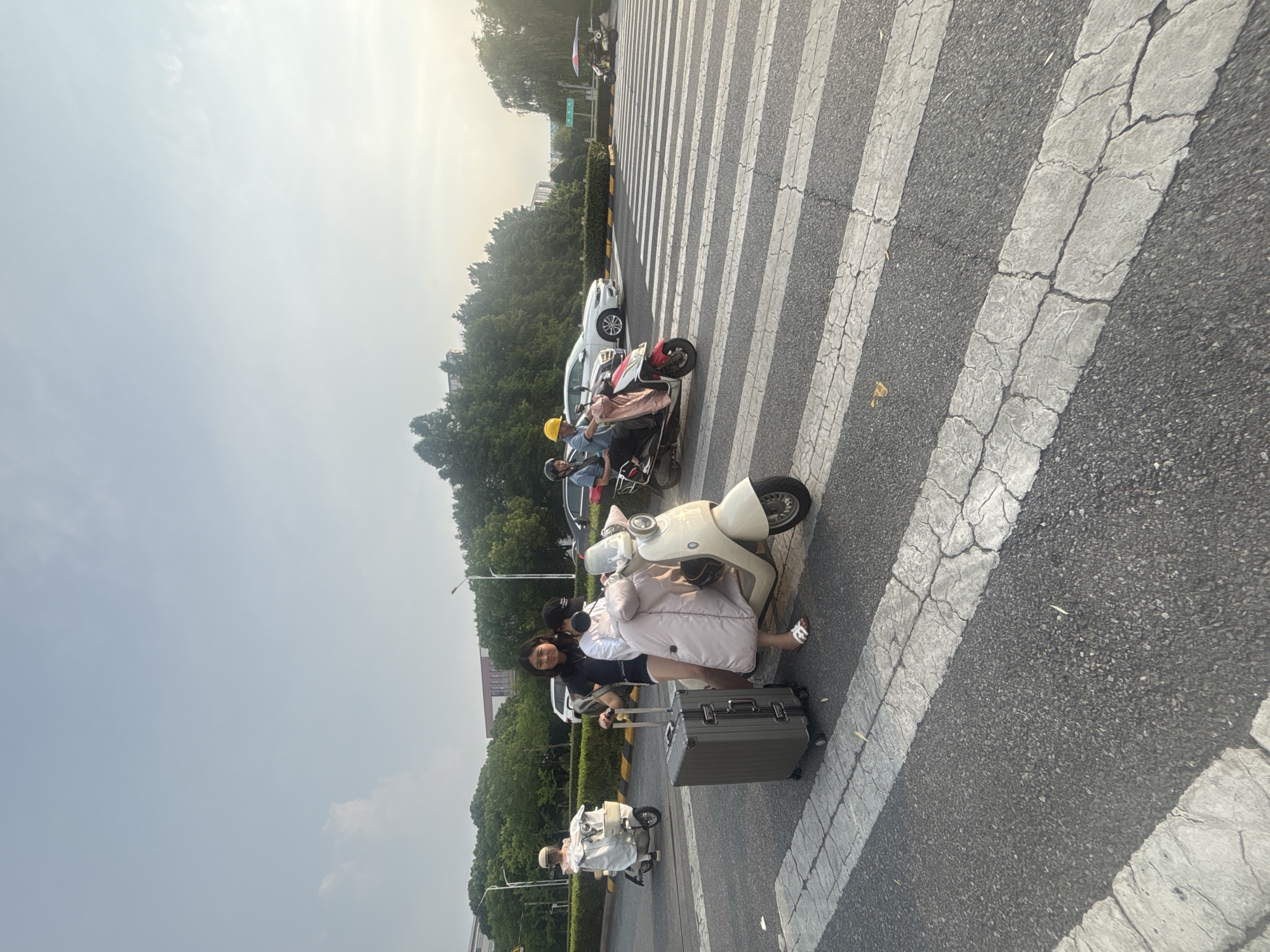 In Suzhou Industrial Park, China, on a street with a crosswalk, two young women are stopped at a pedestrian crossing. The woman in front, wearing a pink coat and white sandals, is seated on a beige scooter. Beside her, the woman behind her, wearing a black t-shirt and shorts, stands holding the handlebars and a gray suitcase. Another scooter, ridden by a person wearing a yellow helmet and a pink garment, is also stopped at the crosswalk. Further down the street, another scooter with two occupants is moving across the intersection, and a white car is visible behind it. The sky is bright, suggesting it is daytime, with some trees and buildings in the background.