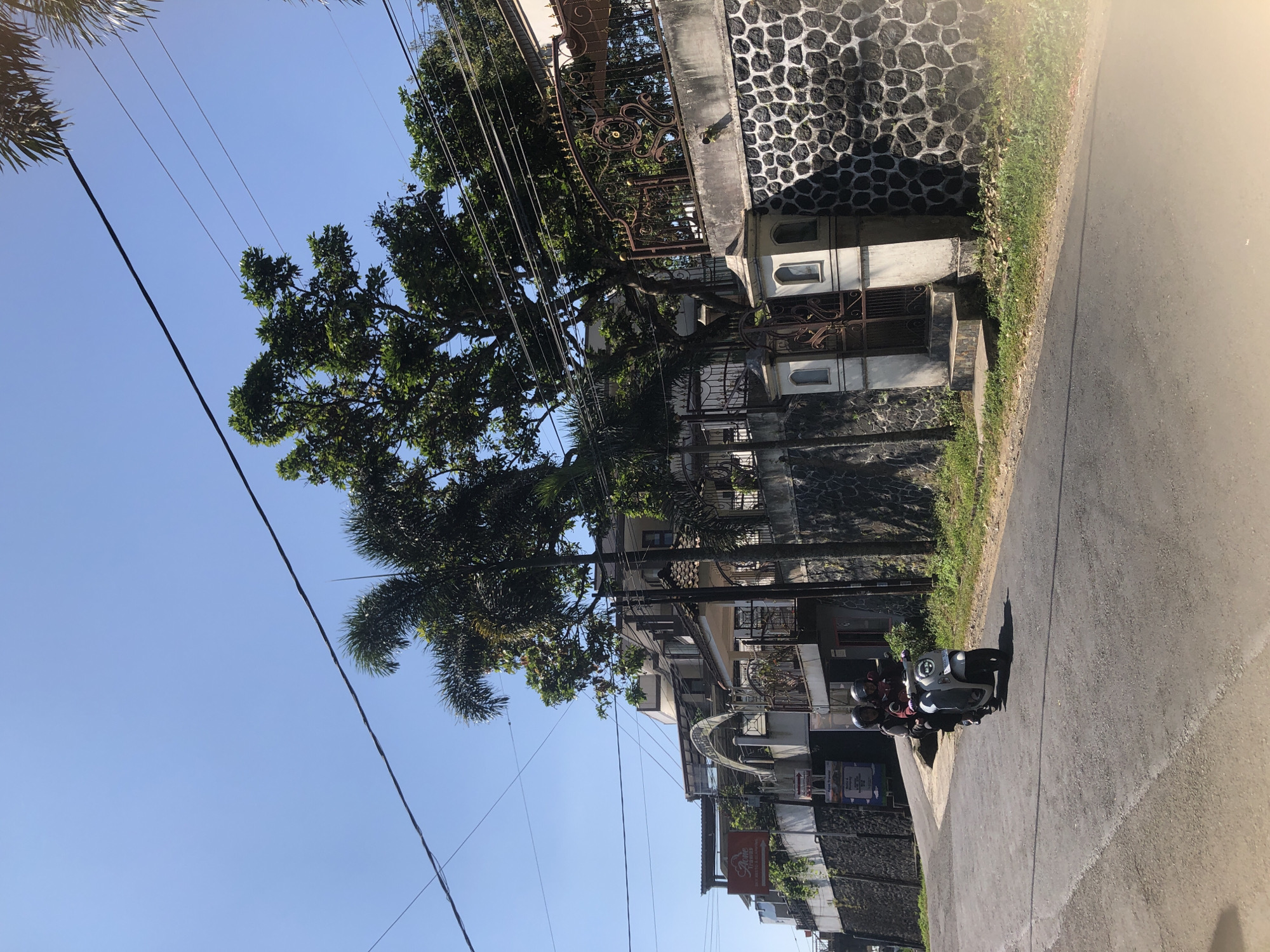 In the city of Trawas, Indonesia, on a clear day, two individuals are riding a scooter down a paved road that curves to the left. The scooter is light gray, and both riders are wearing helmets. The road is bordered by a dry, grassy embankment on the right, which leads up to a stone wall topped with an ornate metal fence. Behind the fence, a multi-story building with white and cream colored walls and a red-tiled roof is visible. Trees with lush green foliage, including palm trees, are scattered throughout the scene, with power lines crisscrossing the bright blue sky above. To the left of the road, another building with a white arched structure and a red sign is partially obscured by trees and vegetation. The stone wall on the right features a white pillar with arched openings and a decorative metal gate.