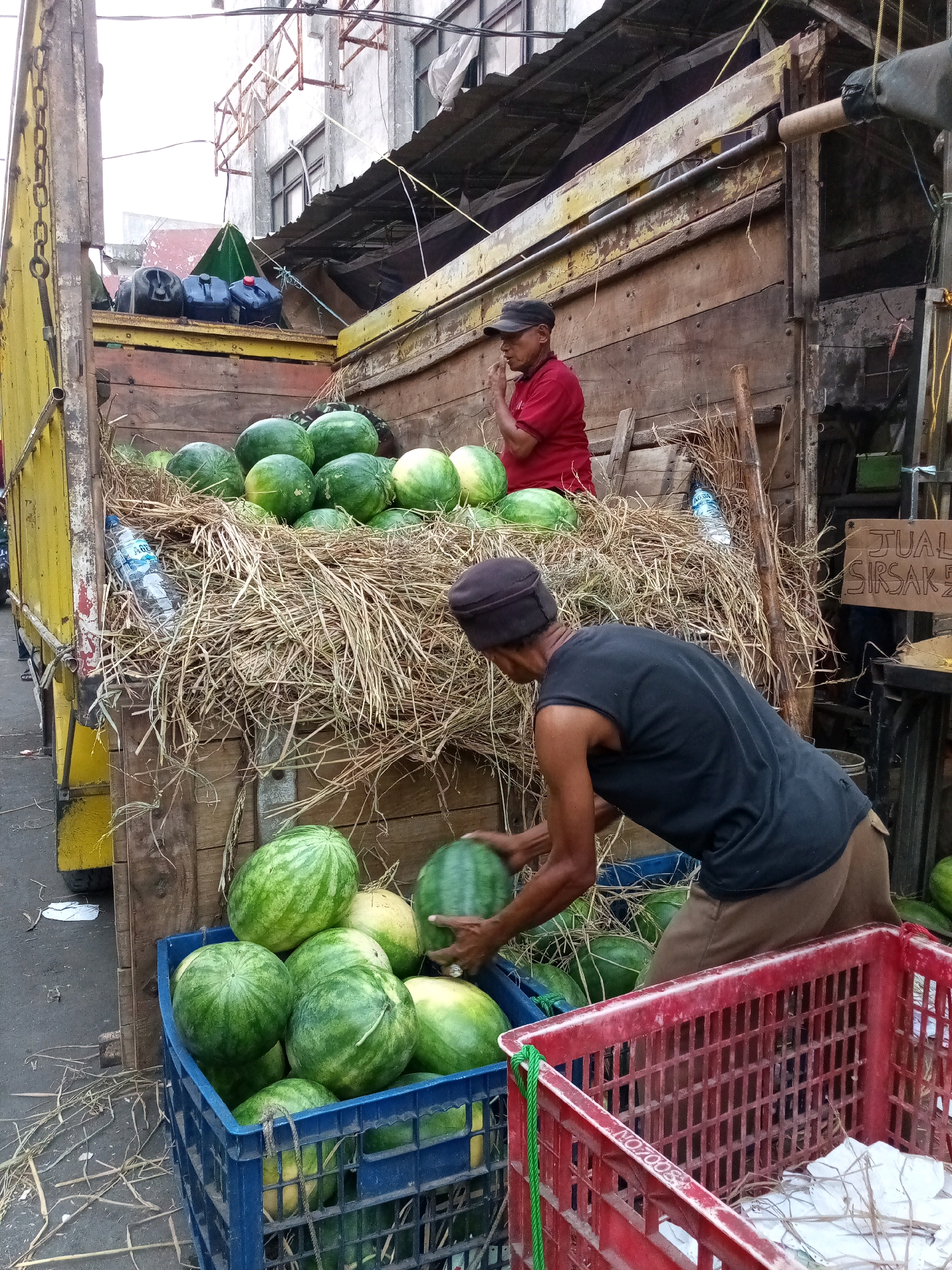 In an outdoor market stall in Bekasi, Indonesia, a man wearing a black t-shirt and brown pants is actively engaged in sorting and placing watermelons into a blue plastic crate. He is positioned in front of a large wooden truck bed filled with hay and more watermelons, some of which are still inside the truck, piled high. Another man, wearing a red shirt and a cap, stands within the truck bed, seemingly observing or assisting. Several plastic water bottles and a green tarp are also visible within the truck. In the foreground, a red plastic crate contains bundled items wrapped in white paper and hay. The overall scene depicts a bustling market environment with a focus on fruit sales.