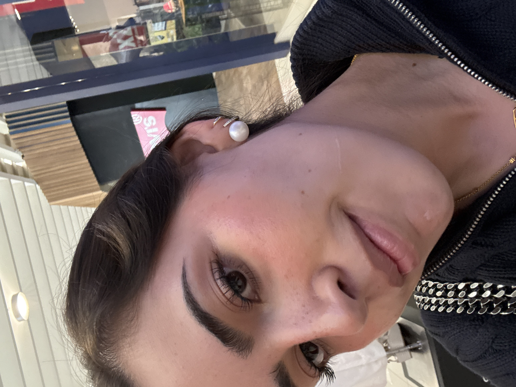 A close-up, angled selfie of a woman with brown hair and defined eyebrows. She has dark eyes with long eyelashes. Her skin tone appears fair with some visible freckles. She is wearing a pearl earring with a sparkling band. She is also wearing a dark, possibly corduroy jacket with a zipper and chain detail. The background shows a blurred reflection in a window of what appears to be a store or commercial building. The location is not provided in the original description, and it is not possible to determine the location in the city of San Francisco Ocotlán (Ocotlán), Mexico from the image alone.