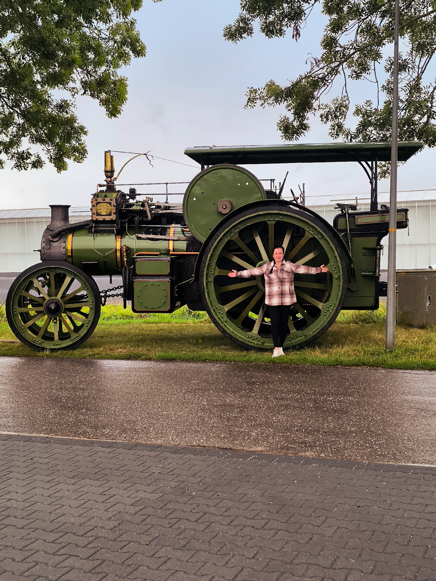 A woman stands in front of a large green steam engine with her arms outstretched. The engine is parked on a grassy area next to a paved road. The woman is wearing a plaid shirt and black pants. In the background, there is a building with a white facade and a tree with green leaves. The sky is overcast. The scene appears to be in Almere, Netherlands.