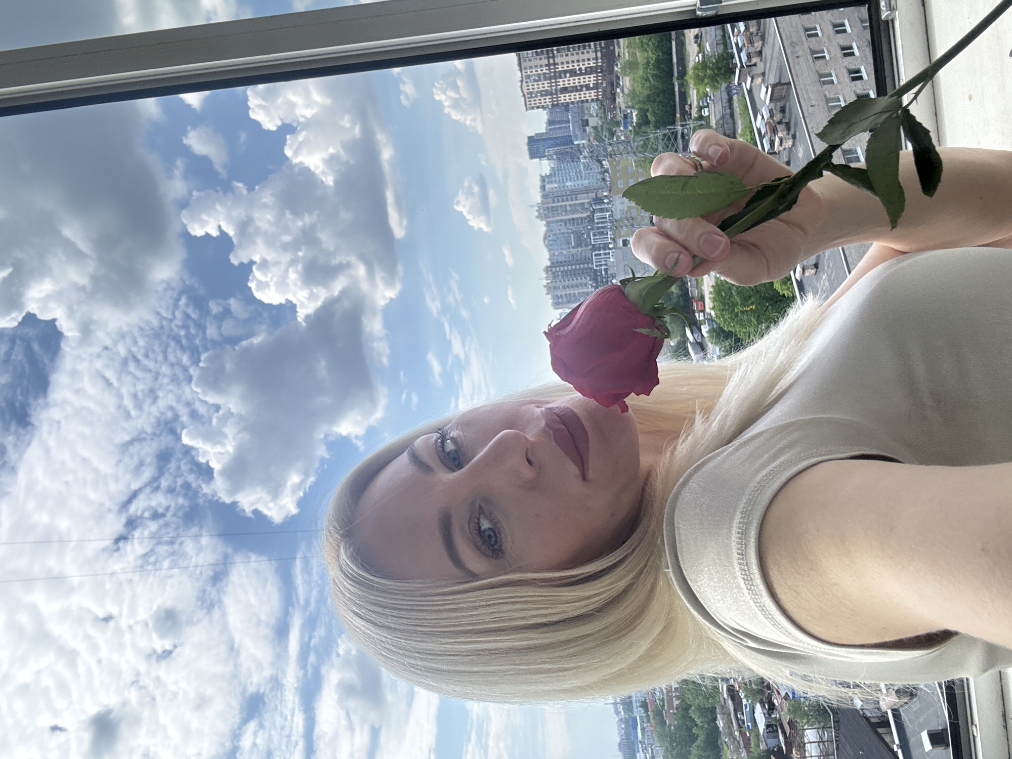 A blonde woman with light eyes and red lipstick is the central subject of the image, holding a single red rose close to her lips. She is wearing a light-colored, possibly beige, short-sleeved top. Her gaze is directed upwards towards the viewer, with a calm and serene expression.
The scene is captured indoors, looking out through a window, a white frame of which is visible at the top and right edge. Beyond the window, a bright blue sky is filled with numerous white, fluffy cumulus clouds, indicating a clear and sunny day. Below the sky, a sprawling urban landscape unfolds. Multiple multi-story buildings of varying architectural styles are visible, forming a dense city skyline. Patches of green foliage from trees are interspersed throughout the urban setting.
The woman appears to be posing for the photograph, delicately holding the rose. The clear sky and bright light suggest it is daytime, likely mid-day. The prominent buildings and urban density indicate a city environment. No visible text is present in the image. The context suggests the location is an unknown city in Russia.